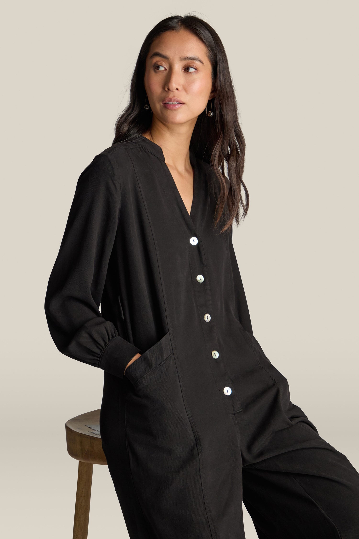 A woman with long dark hair, wearing a black Fluid Twill Jumpsuit that features a modern rounded neckline, sits on a stool with her hands in her pockets.