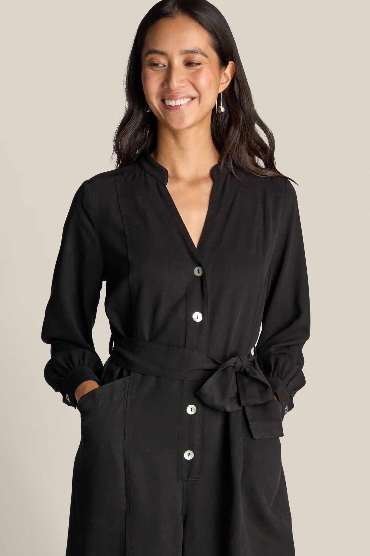 A woman with long dark hair is wearing the Fluid Twill Jumpsuit in black, which features a tied waist. She is facing forward, smiling, and looks effortlessly chic in this contemporary outfit.