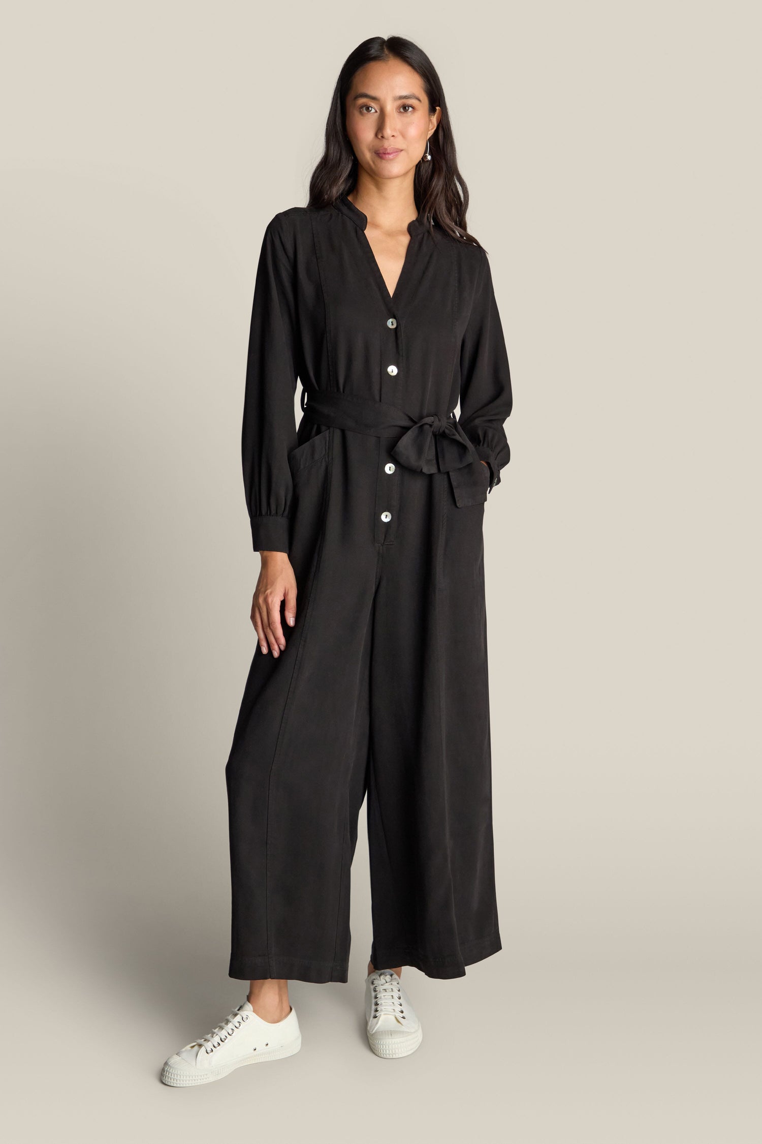 A person wearing the black Fluid Twill Jumpsuit, featuring white buttons and pockets, along with white sneakers, stands against a plain background. The contemporary rounded neckline adds a modern touch to this eco-friendly fabric ensemble.