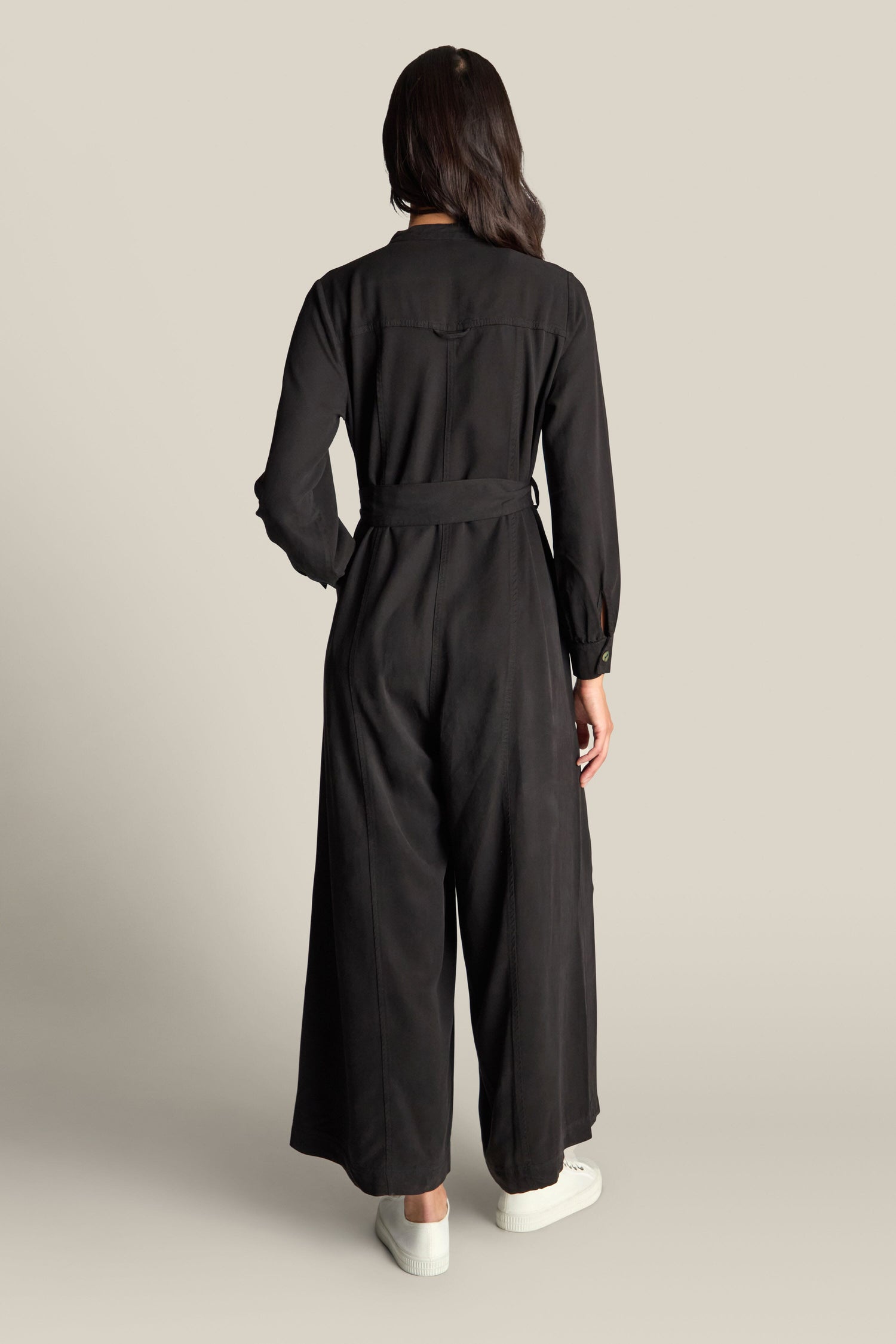 A person with long hair stands facing away, donning a Fluid Twill Jumpsuit crafted from eco-friendly fabric with a modern rounded neckline, paired with white sneakers.