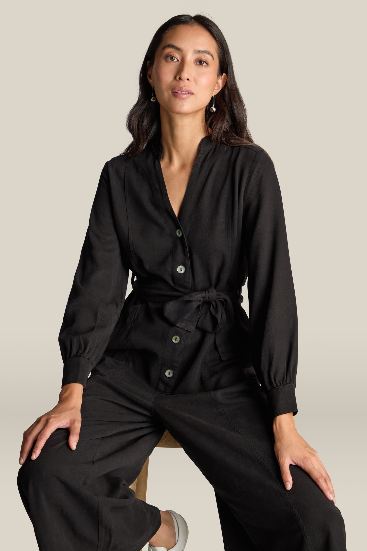 A woman with long dark hair sits on a stool, wearing the Fluid Twill Jumpsuit made from eco-friendly fabric, against a plain background.