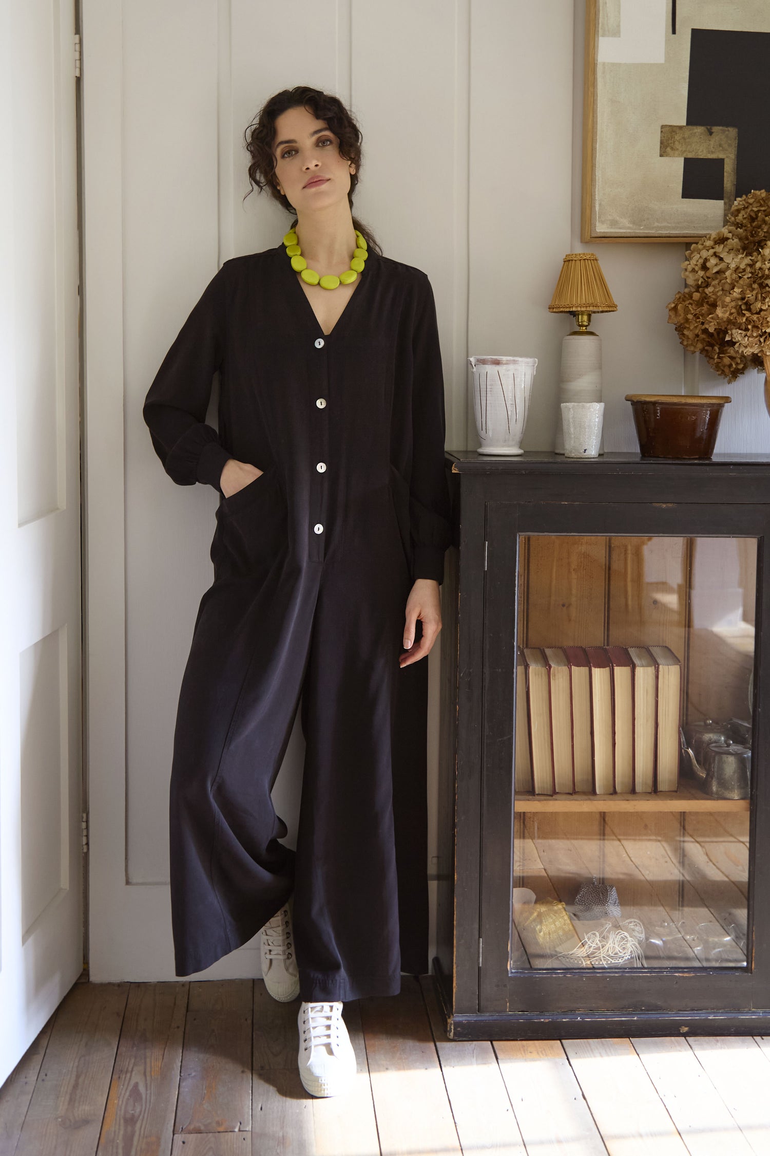 A woman in a relaxed-fit Fluid Twill Jumpsuit, crafted from eco-friendly fabric, stands next to a glass-fronted cabinet filled with books and decorative items. She pairs her outfit with white sneakers and a yellow necklace.