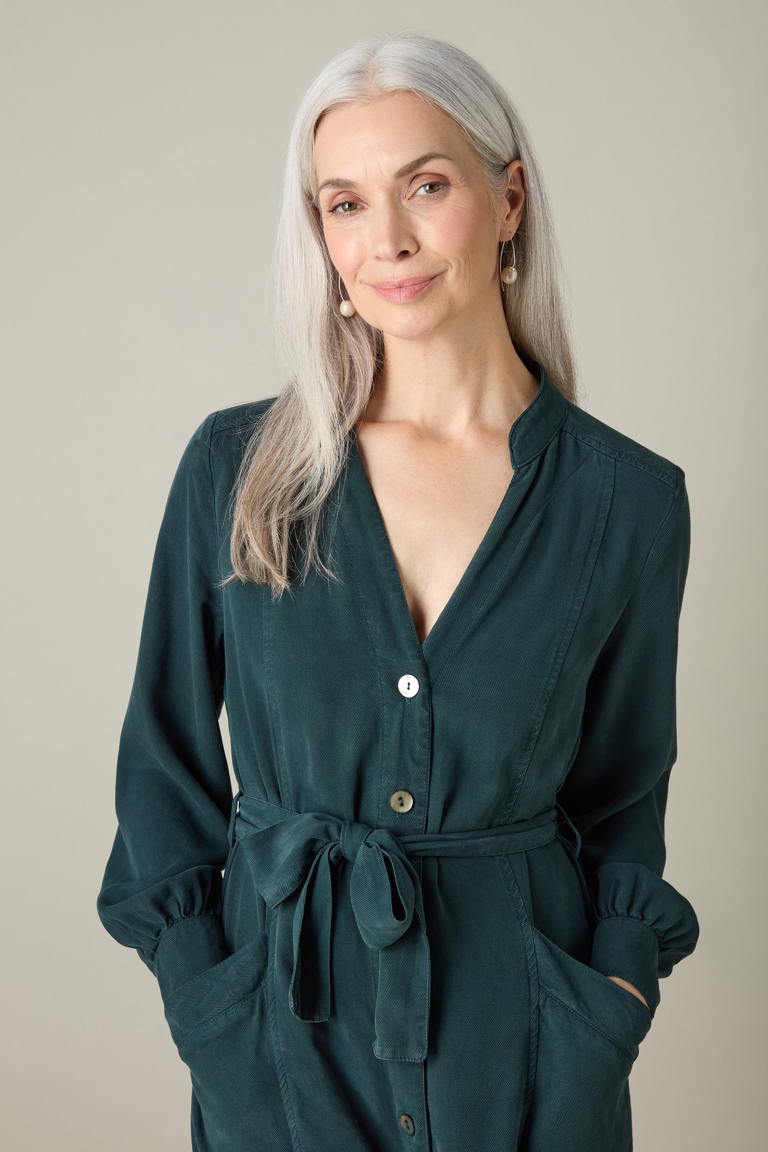 A woman with long gray hair, dressed in a dark green Fluid Twill Jumpsuit featuring a modern rounded neckline and accessorized with earrings, stands with her hands in her pockets and offers a subtle smile.