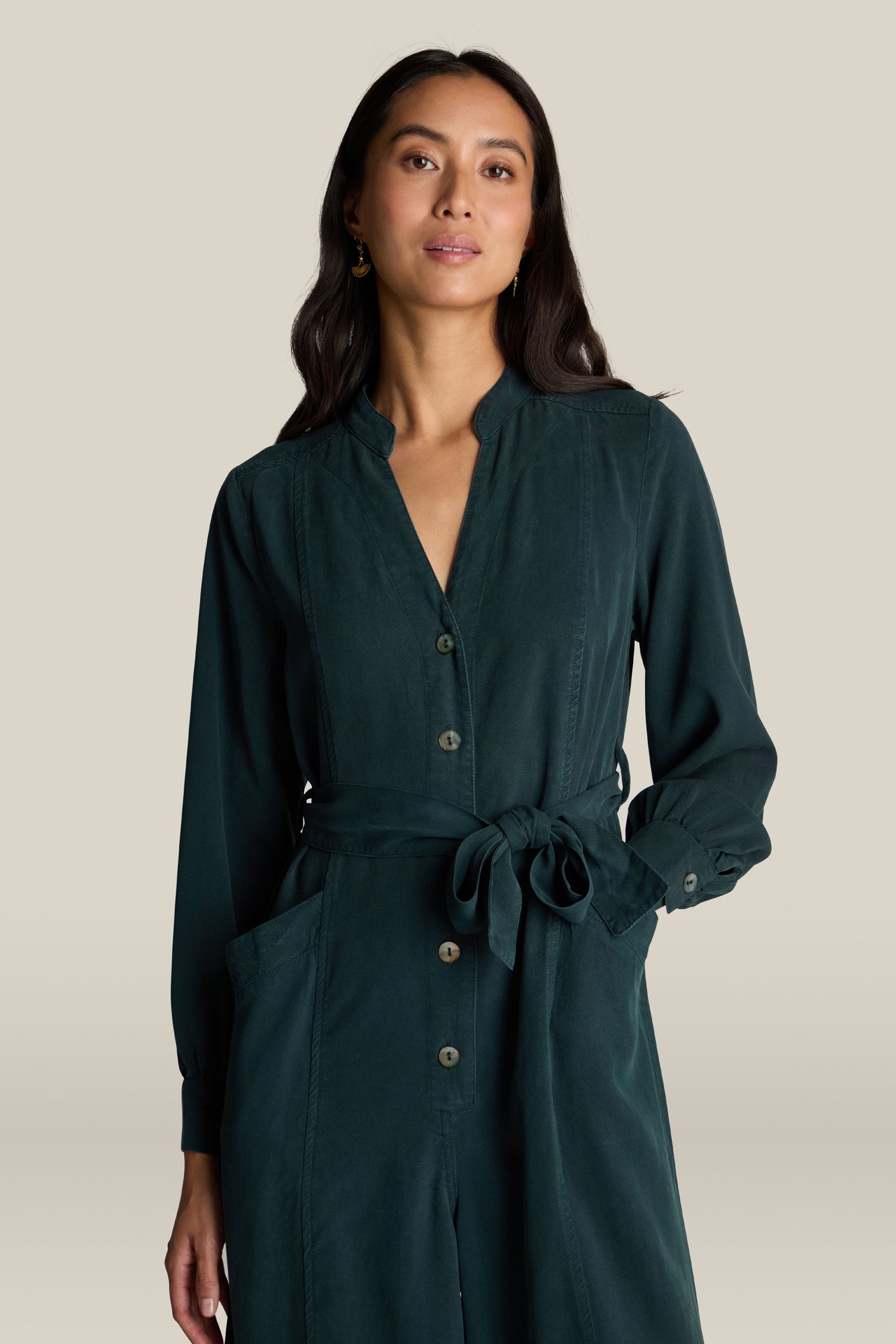 A woman with long dark hair, wearing a dark green Fluid Twill Jumpsuit made from eco-friendly fabric, stands against a light background, hands in pockets, and looking forward.