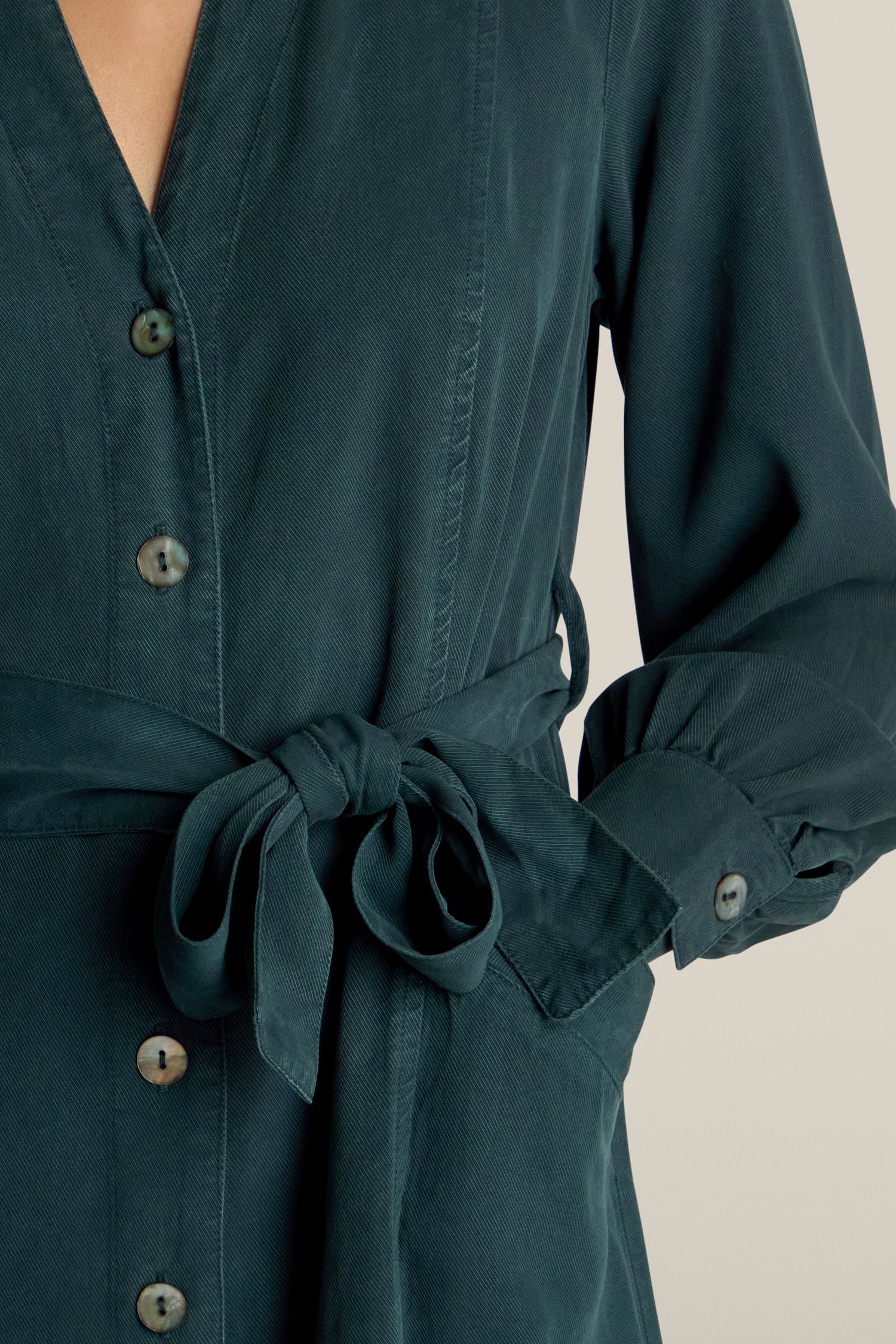 Close-up of a person wearing a dark green Fluid Twill Jumpsuit with a tied belt at the waist, showing buttons and partially rolled-up sleeves.