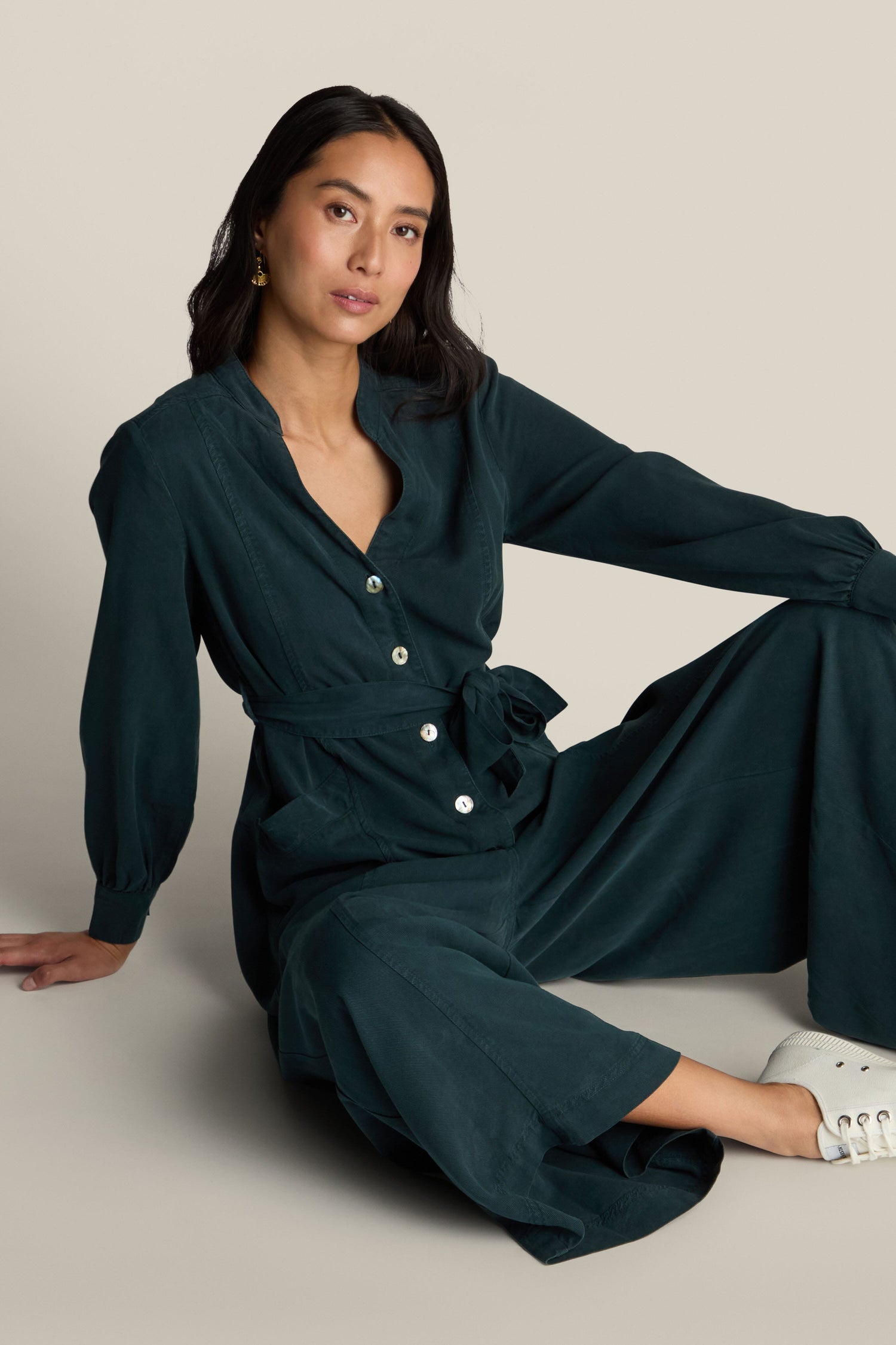 A woman, seated on the floor, wears a dark green Fluid Twill Jumpsuit that features a contemporary rounded neckline and a waist tie. One hand is placed on the ground while the other rests on her knee. She has long hair and gazes at the camera.