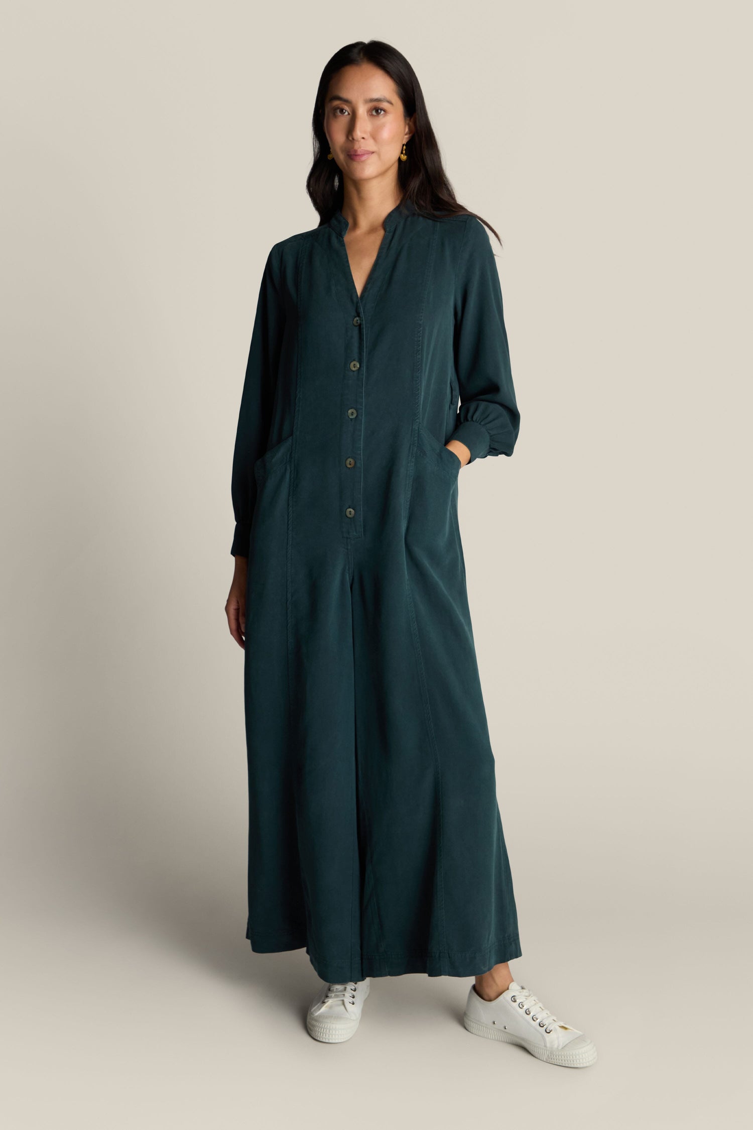 A woman stands against a plain background wearing a Fluid Twill Jumpsuit in dark green, crafted from eco-friendly fabric, paired with white sneakers. She has her hands in the jumpsuit pockets.