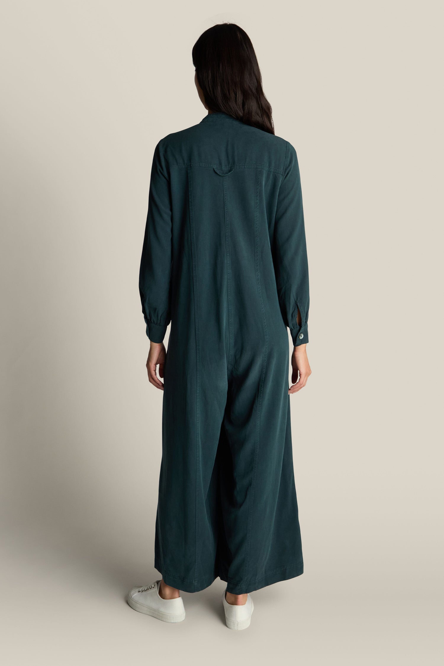 A person with dark hair stands facing away from the camera, wearing the Fluid Twill Jumpsuit in a long-sleeved, dark teal design featuring a contemporary rounded neckline and white sneakers, against a plain background.