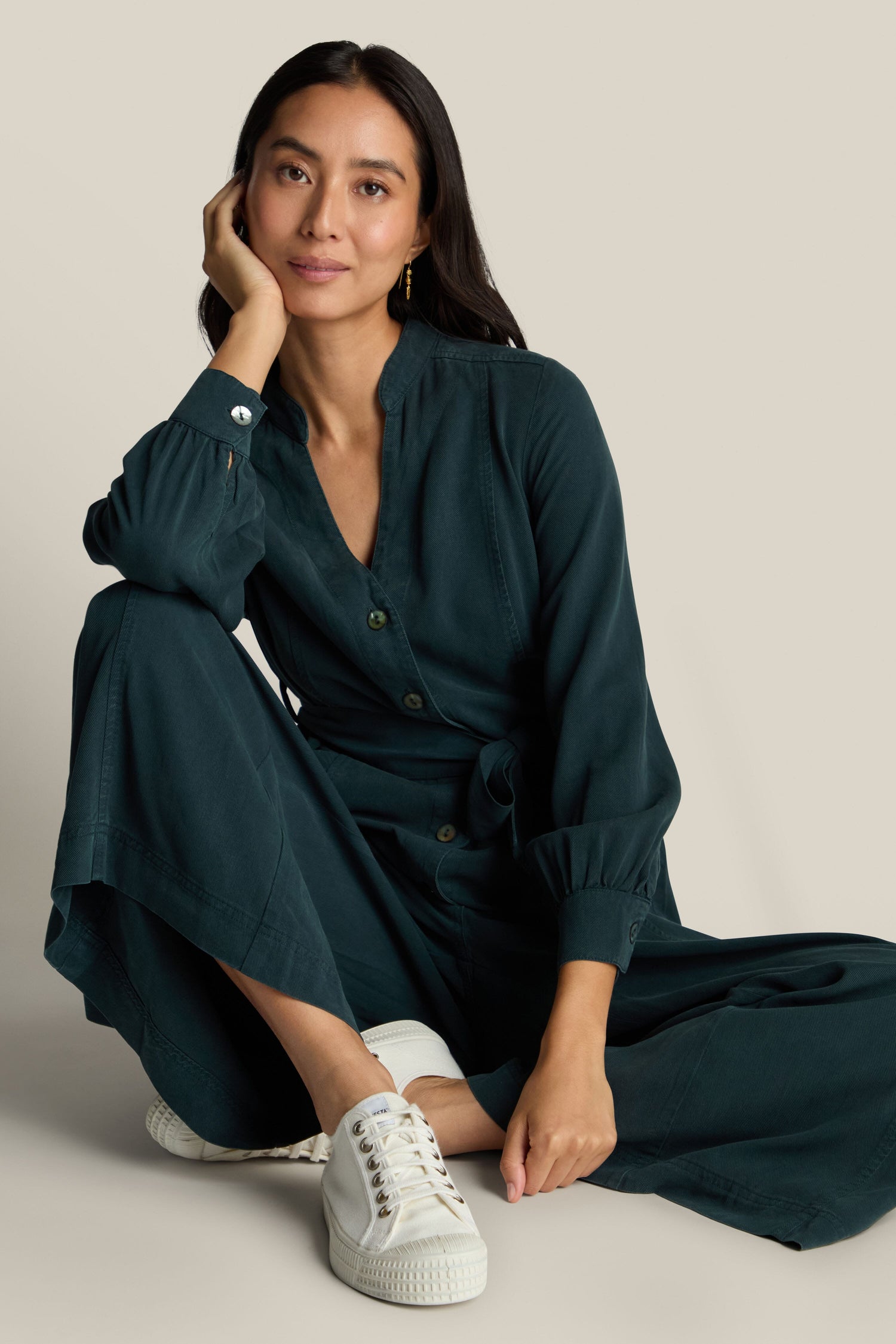 A person with long dark hair is sitting on the floor, dressed in a dark green Fluid Twill Jumpsuit crafted from eco-friendly fabric and white sneakers. They have one hand resting on their chin and a calm expression.