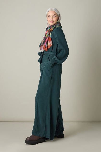 A woman with gray hair is shown in profile, adorned in a Fluid Twill Jumpsuit created from eco-friendly fabric, complemented by a colorful scarf and black boots against a simple light-colored background.