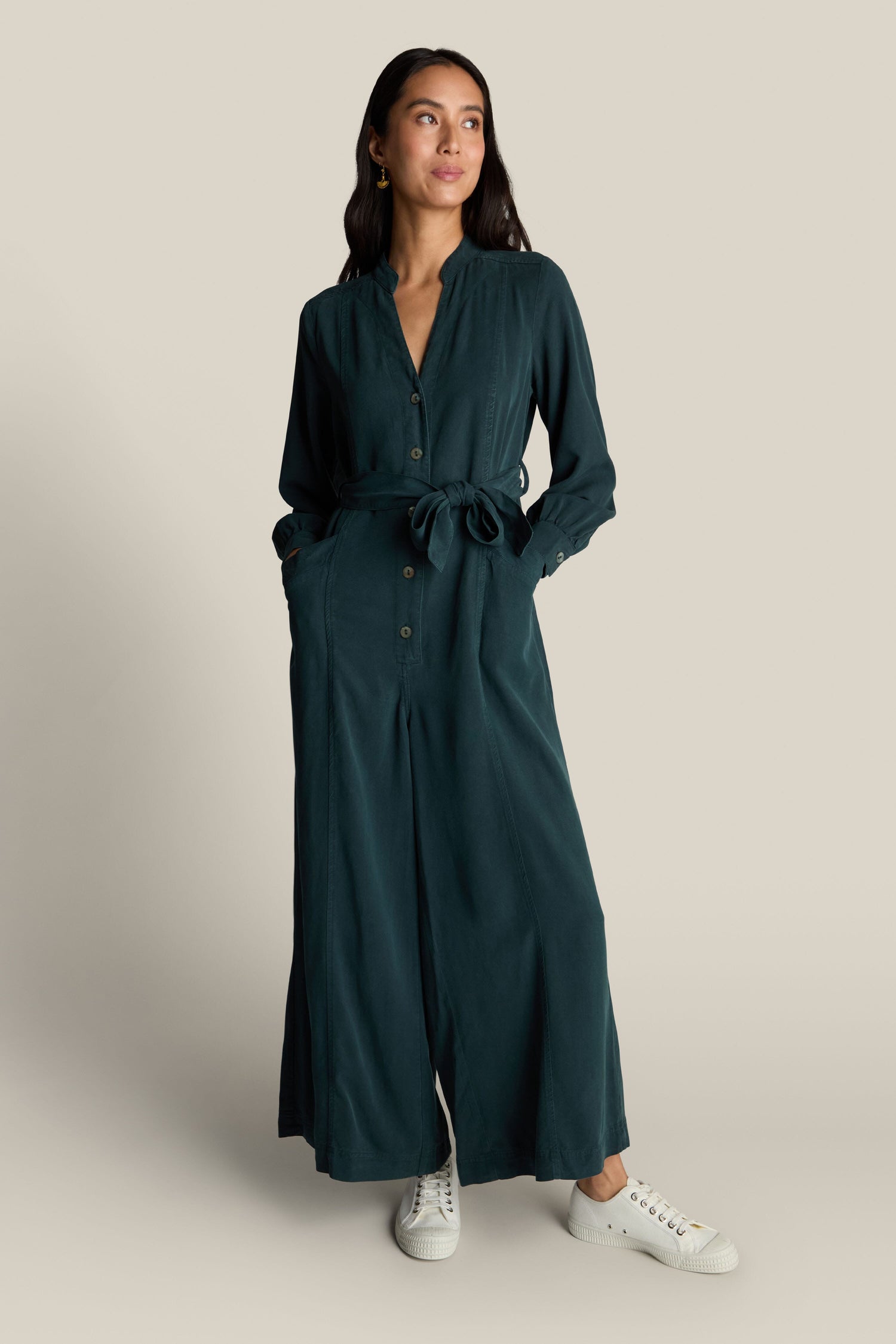 A woman stands wearing a dark green, long-sleeve Fluid Twill Jumpsuit with a tied waist and contemporary rounded neckline, crafted from eco-friendly fabric, paired with white sneakers.