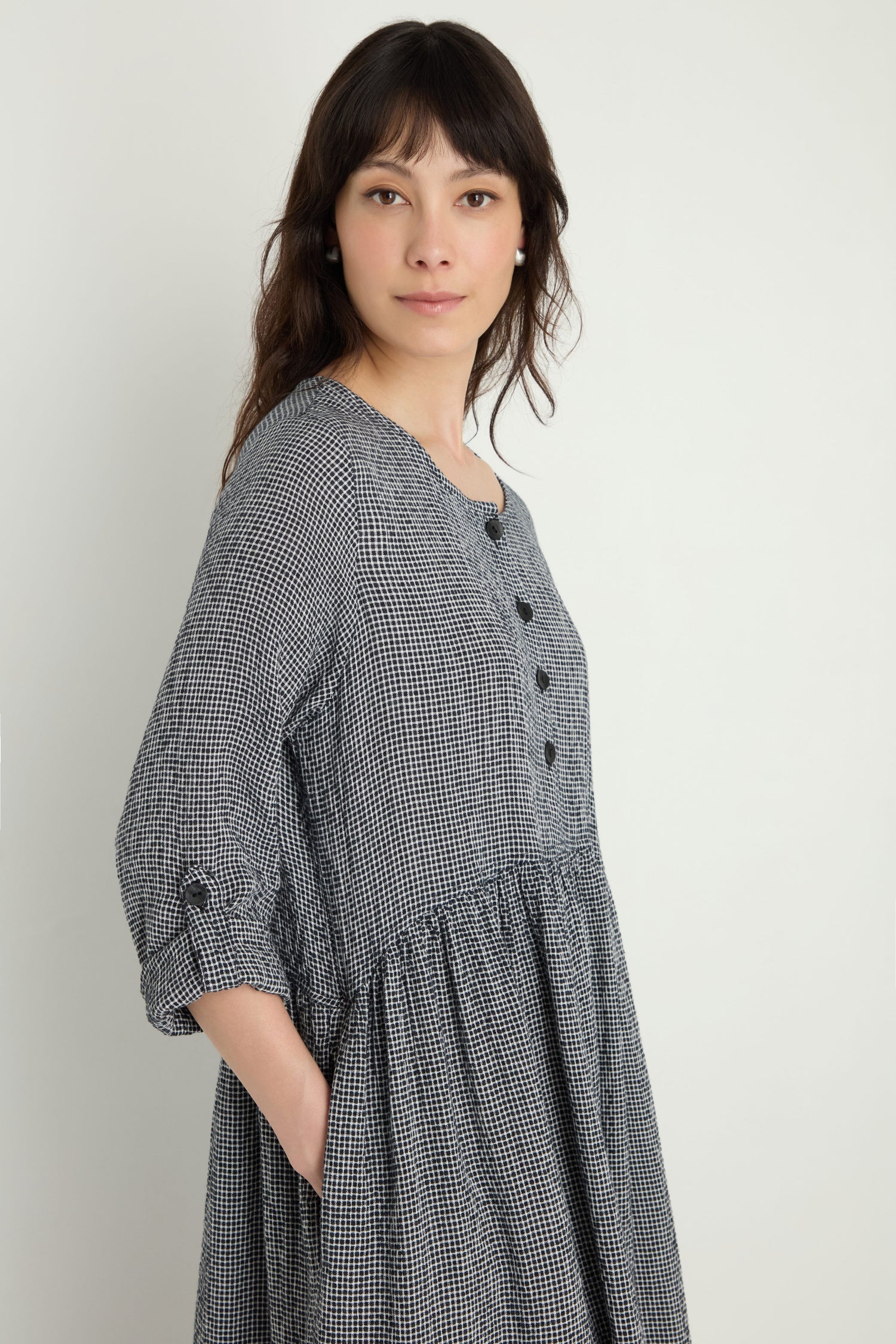 A woman stands against a plain light background wearing the Micro Gingham Bubble Dress (MTD6166-MGI), a loose-fitting, long-sleeve, black-and-white checkered button dress.