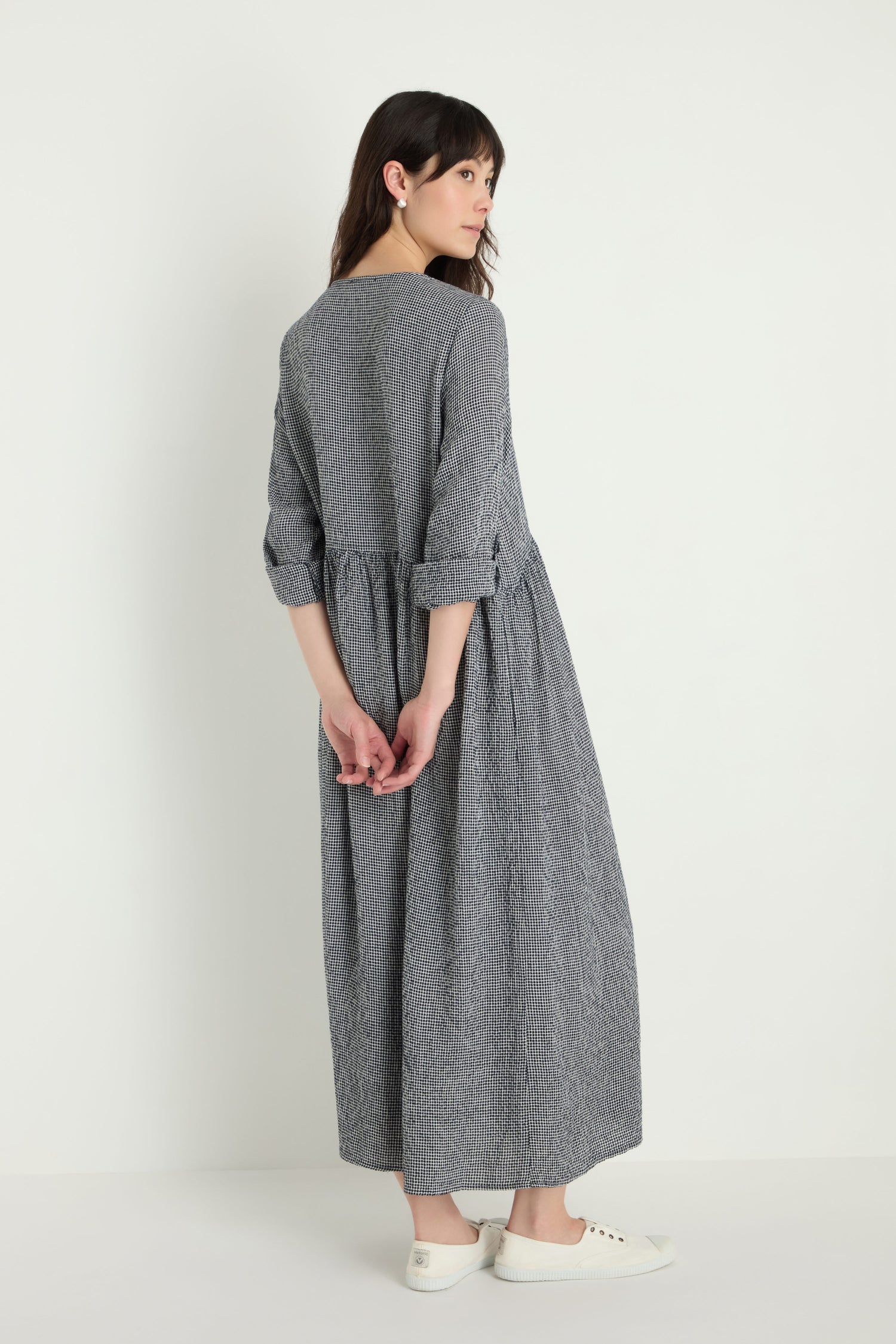 A woman stands against a white background wearing the Micro Gingham Bubble Dress, a long, loose-fitting gray dress with rolled-up sleeves and white sneakers. She is facing away with her hands clasped behind her back.