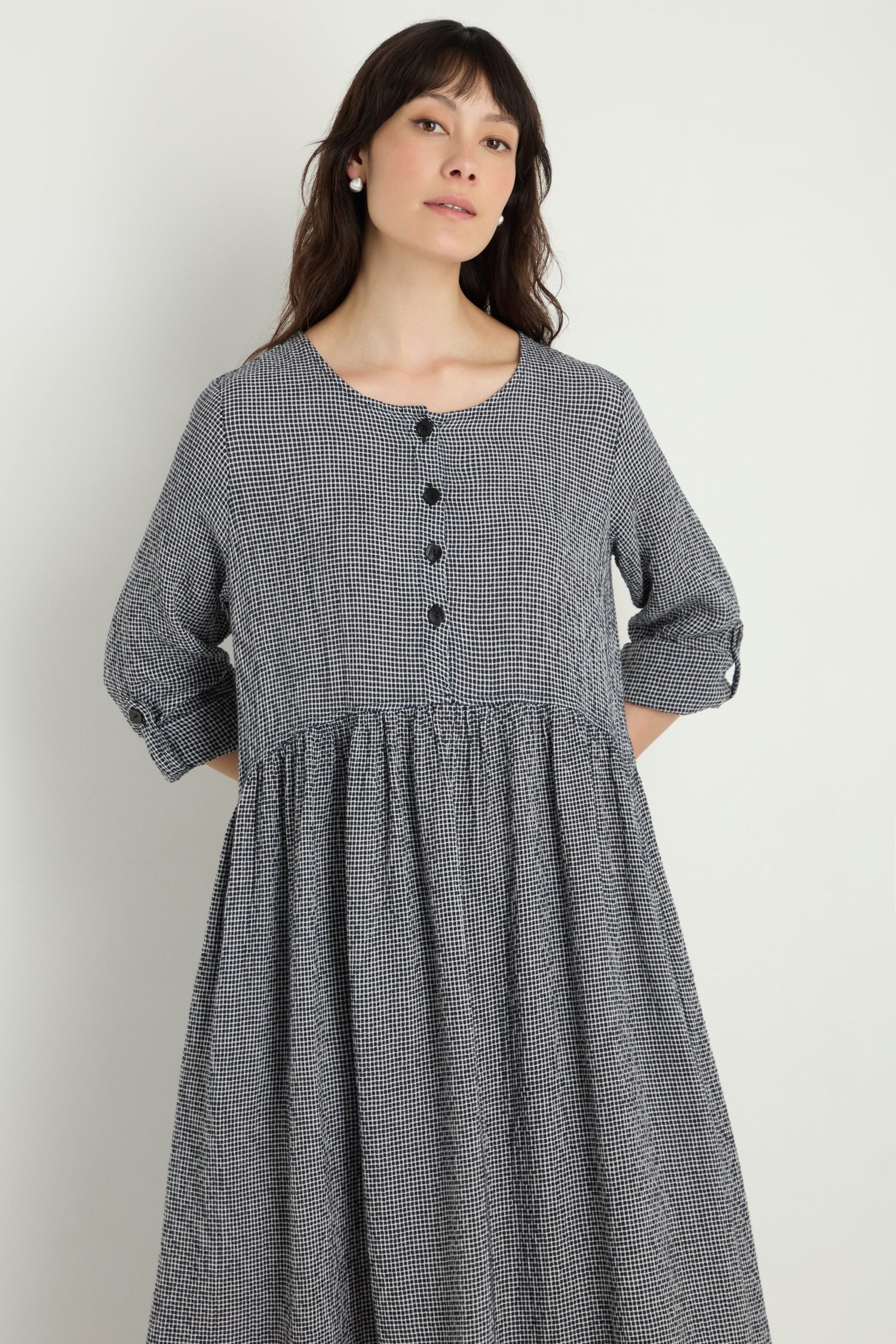 A woman stands against a plain light background wearing the Micro Gingham Bubble Dress, a loose-fitting, long-sleeve black and white checkered dress with buttons on the upper front.