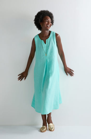 Smiling woman stands by a white wall in the Linen Notch Neck Sleeveless Dress—an aqua A-line silhouette—styled with a gold necklace and woven gold slip-on shoes, making it perfect for your summer wardrobe.