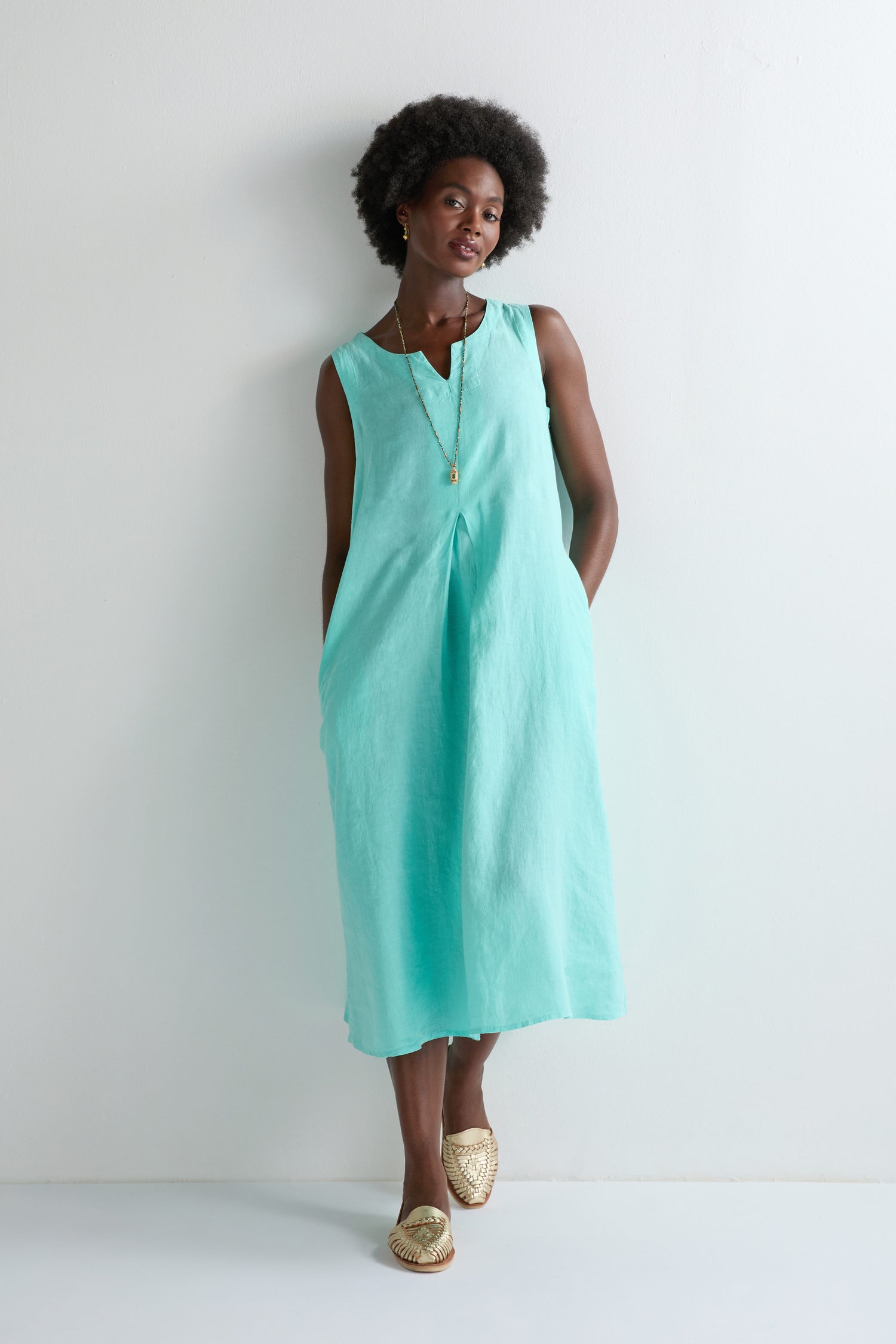 A woman stands against a white wall wearing the Linen Notch Neck Sleeveless Dress in turquoise, styled with a gold necklace and woven slip-on shoes—a chic choice to refresh any summer wardrobe.