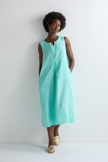 A woman stands against a white wall wearing the Linen Notch Neck Sleeveless Dress in turquoise, styled with a gold necklace and woven slip-on shoes—a chic choice to refresh any summer wardrobe.