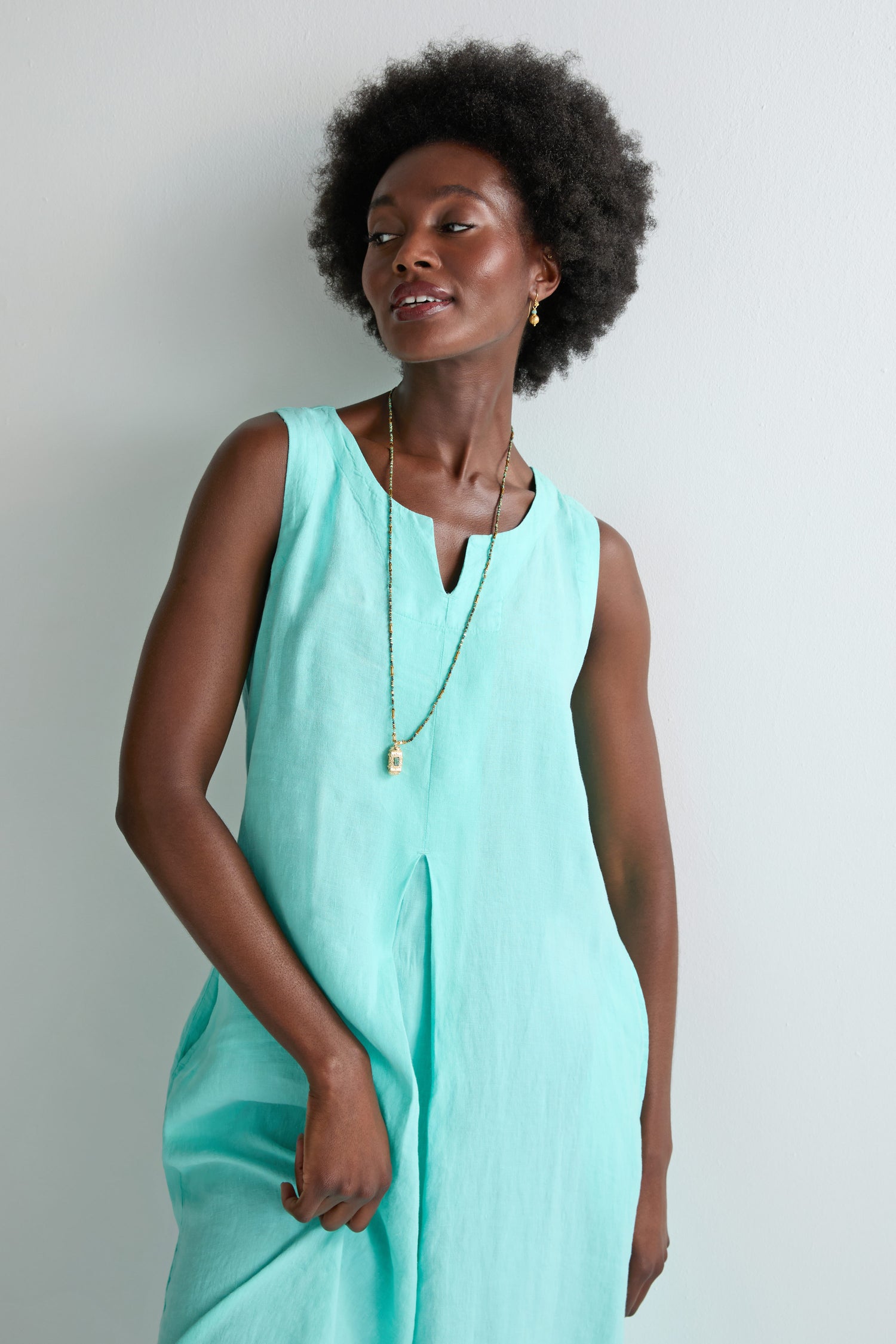 A woman stands against a plain light-colored wall, effortlessly showcasing her summer style in the Linen Notch Neck Sleeveless Dress—a light turquoise, linen piece featuring an A-line silhouette and a notch neckline.