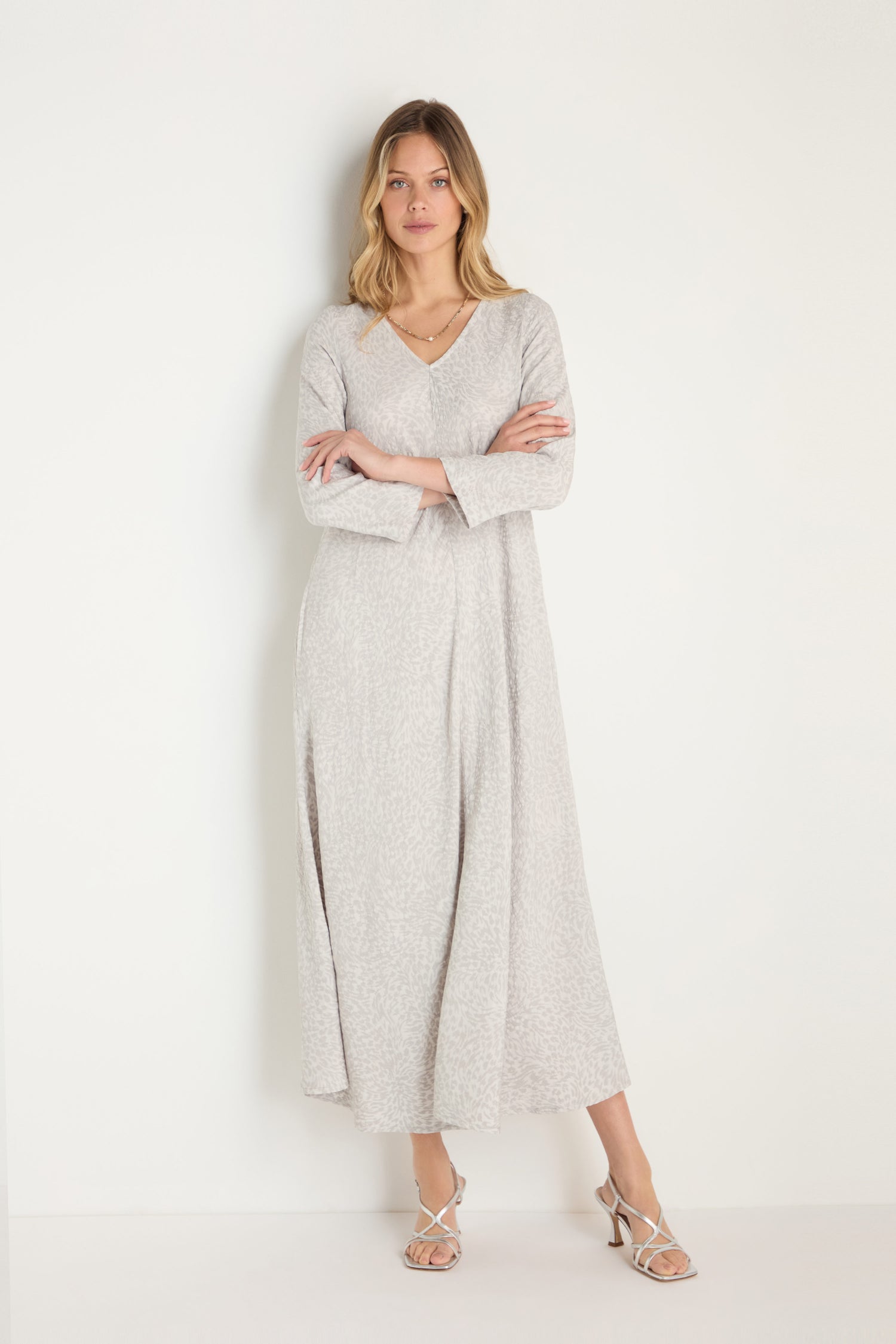 A woman with light hair and a neutral expression stands against a white wall, wearing the Animal Textured Jacquard Maxi Dress in light gray, paired with white strappy heels.