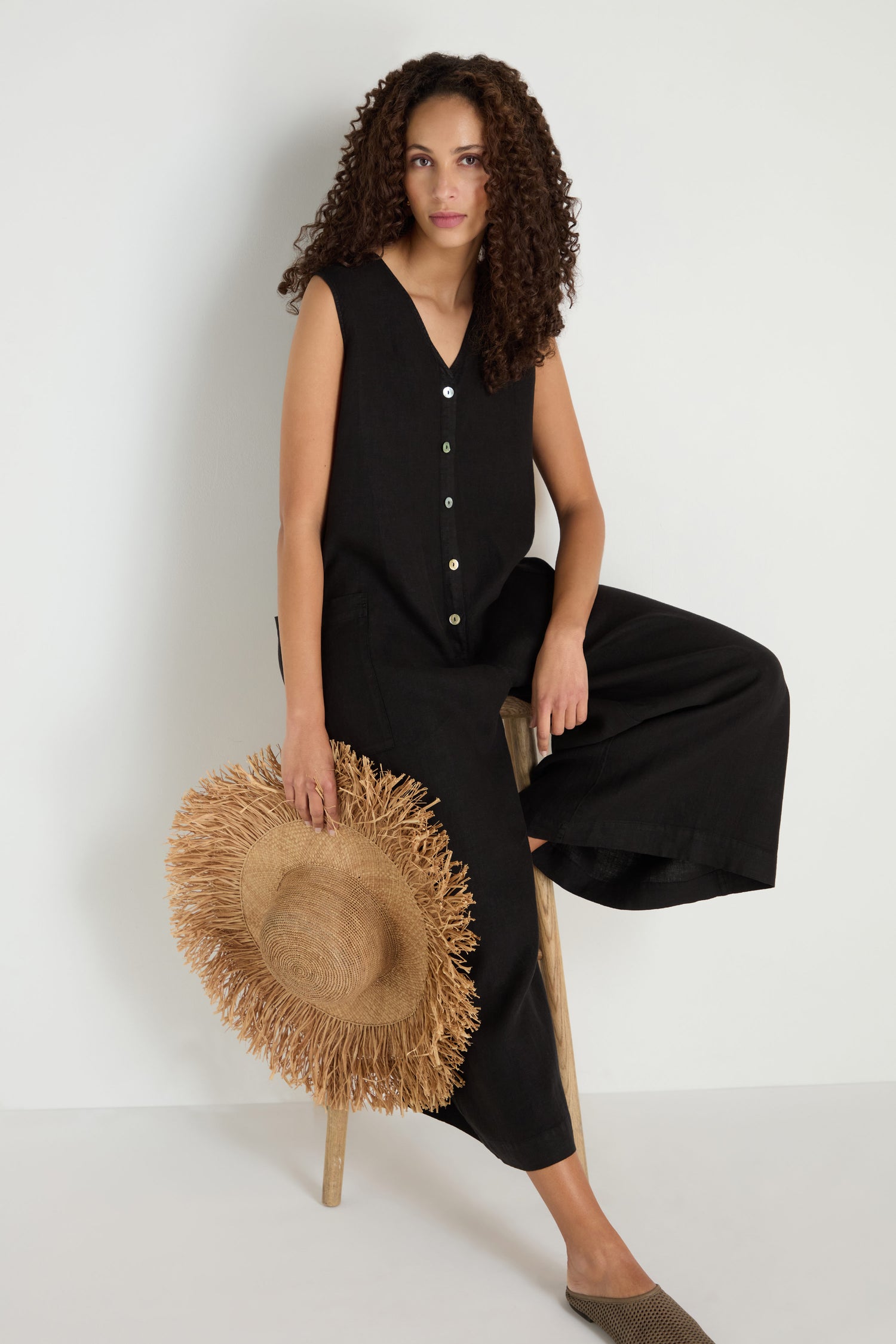 A woman with curly hair sits on a stool against a white background, wearing the Linen Relaxed Jumpsuit with wide legs and sleeveless design, and holding a wide-brimmed straw hat.