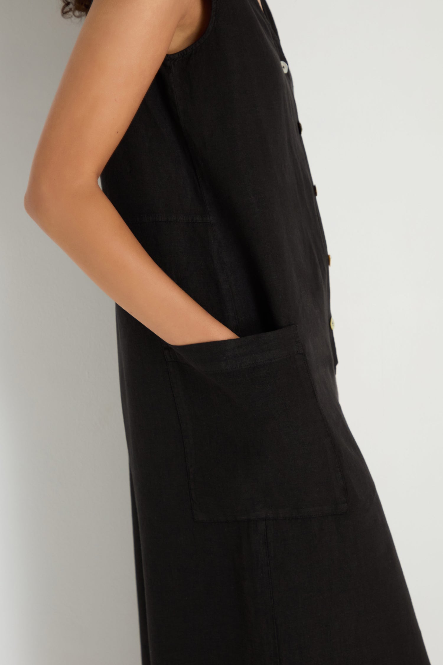 Someone stands against a plain white background wearing the Linen Relaxed Jumpsuit—a black, sleeveless, relaxed fit piece featuring large front pockets.