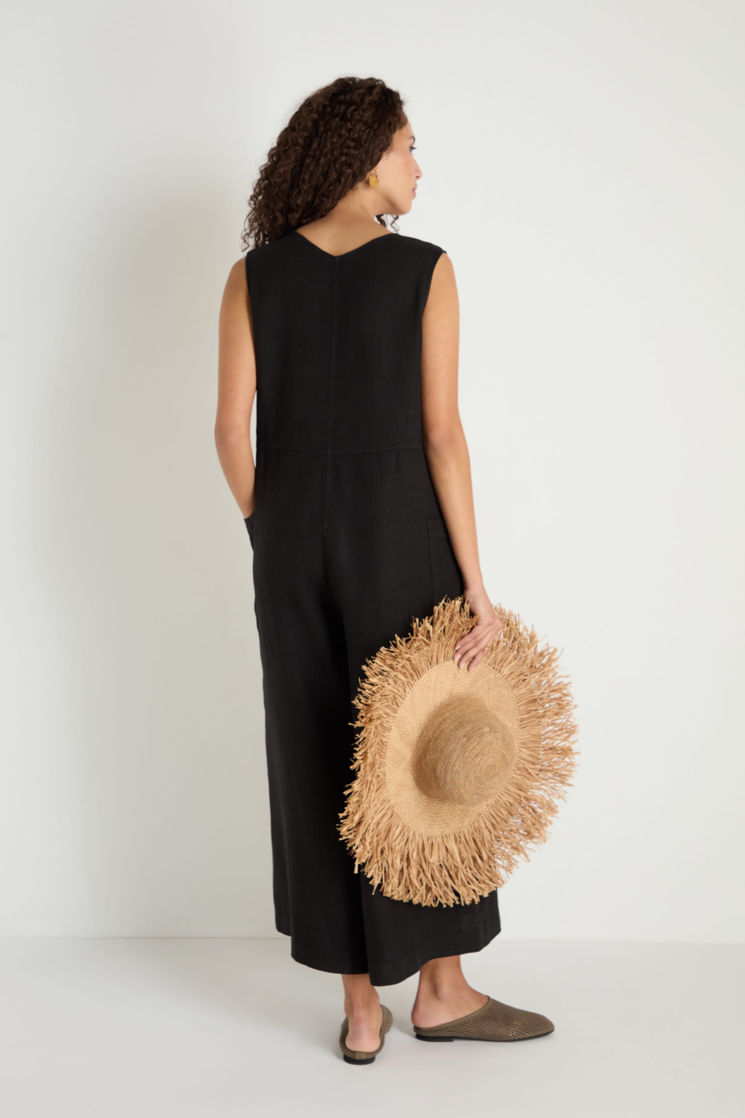A woman faces away in the Linen Relaxed Jumpsuit, a sleeveless black wide-leg linen piece. She holds a straw hat, her curly hair showing, and wears brown slip-on shoes.