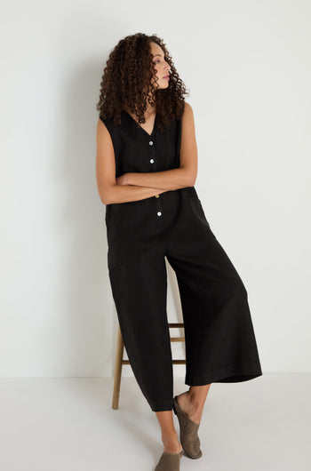 A woman with curly hair sits on a wooden stool, wearing the Linen Relaxed Jumpsuit and taupe slip-on shoes, against a plain white background.