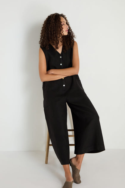 A woman with curly hair sits on a wooden stool, wearing the Linen Relaxed Jumpsuit and taupe slip-on shoes, against a plain white background.