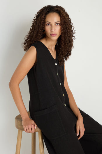 A person with curly hair sits on a wooden stool, wearing the Linen Relaxed Jumpsuit in black with a sleeveless, relaxed fit, facing the camera against a plain light background.