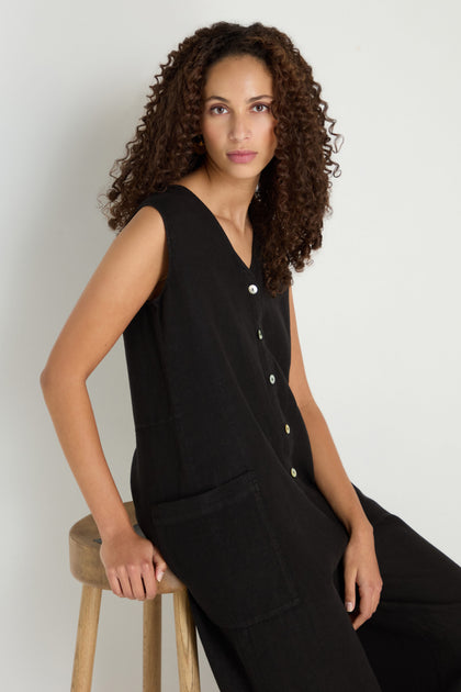 A person with curly hair sits on a wooden stool, wearing the Linen Relaxed Jumpsuit in black with a sleeveless, relaxed fit, facing the camera against a plain light background.