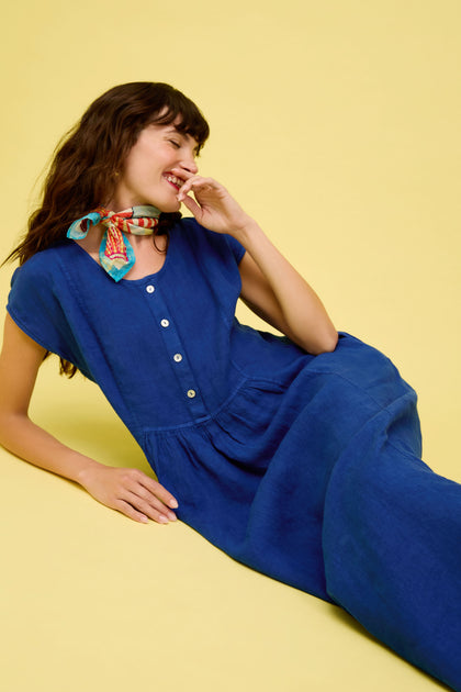 A woman in the Linen Button Cerulean Placket Dress and a colorful scarf reclines on a yellow background, smiling and gazing to the side.