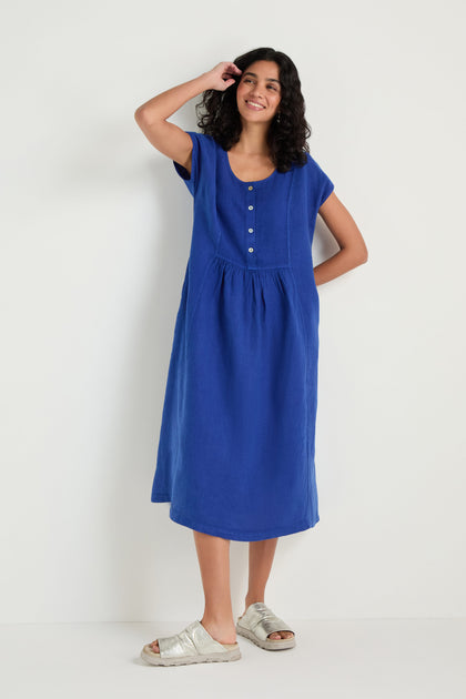 A woman stands against a plain white background, wearing the Linen Button Cerulean Placket Dress styled with silver sandals.