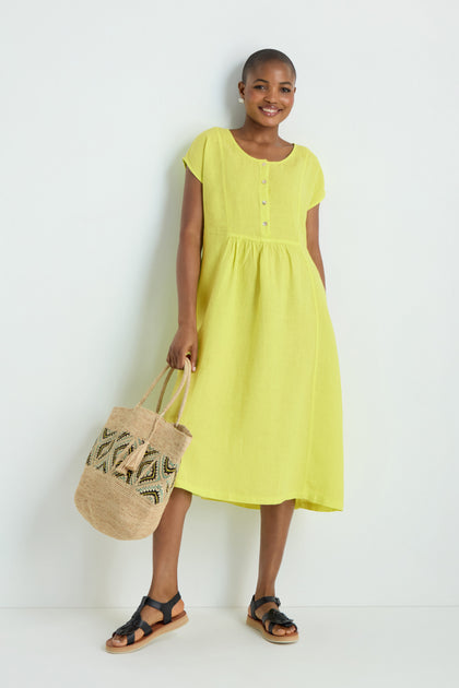 A person wearing the Linen Button Lemon Placket Dress, holding a woven tote bag and black sandals, stands against a plain white background.