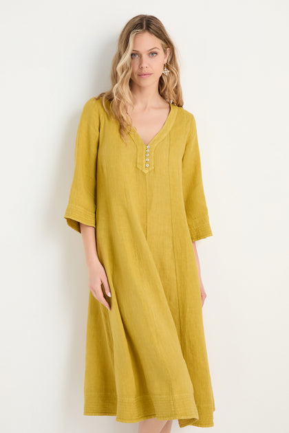 A woman models the Linen Seam Golden Flare Dress, featuring panel seams, three-quarter sleeves, a buttoned V-neck, and a loose yellow flare silhouette, standing against a plain white background.