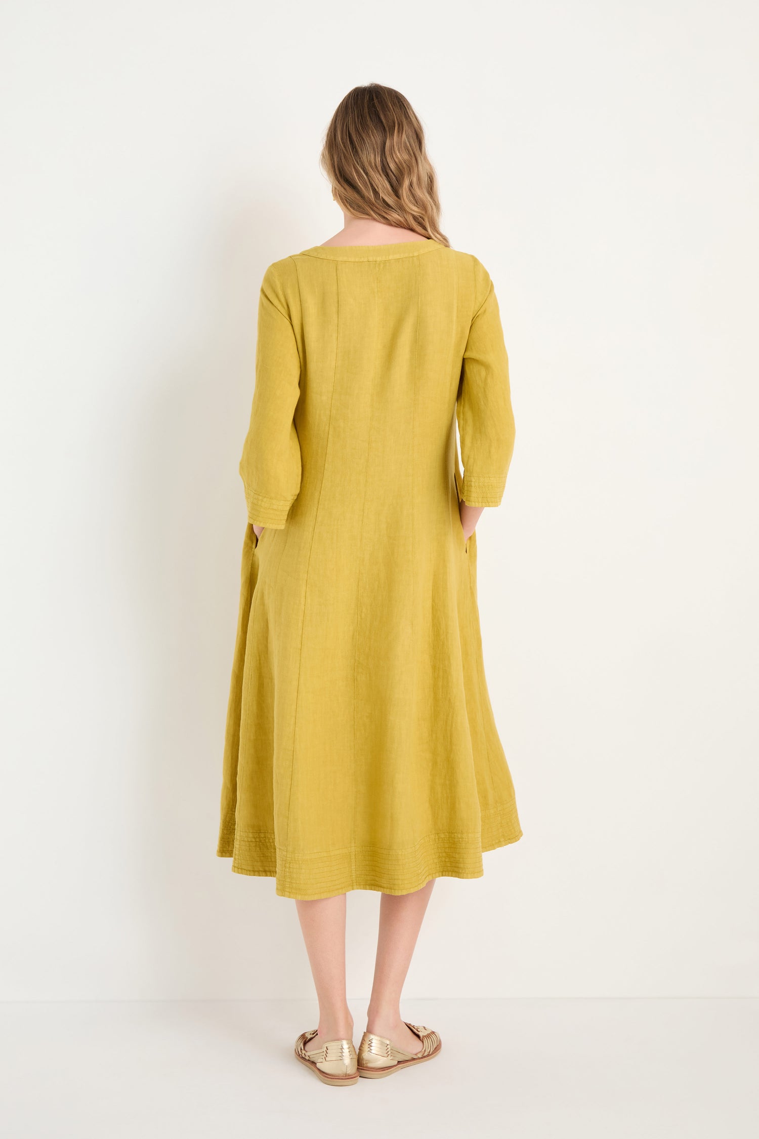 A woman faces away, wearing the Linen Seam Golden Flare Dress—loose-fitting with long sleeves and panel seams—paired with gold sandals against a plain white background.