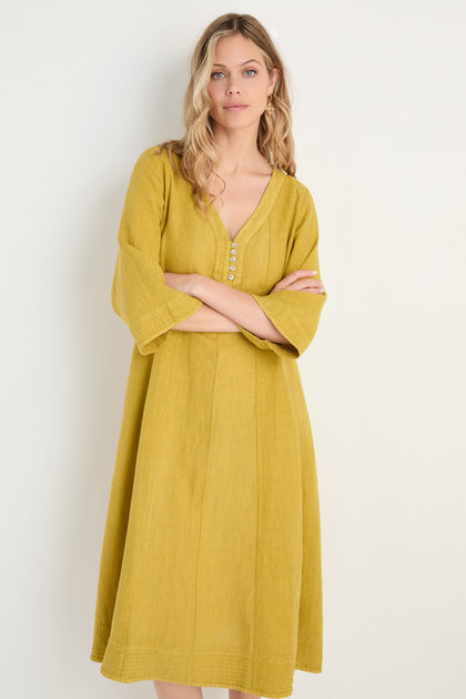 A woman models the Linen Seam Golden Flare Dress in mustard yellow, showcasing its V-neckline, panel seams, and button details against a simple light background.