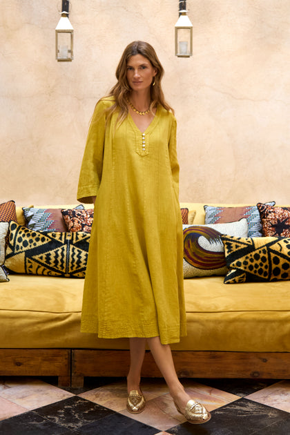 A woman in a Linen Seam Flare Golden Dress and gold shoes stands in front of a patterned sofa with assorted decorative pillows and hanging lanterns above.