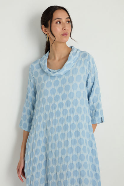 A person wears the Spot Textured Jacquard Bubble Dress, a light blue, long-sleeved dress with large polka dots, standing with one arm bent across their stomach.