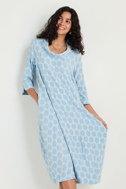 A woman smiles and holds the sides of her Spot Textured Jacquard Bubble Dress, a light blue, midi-length garment with a polka dot pattern, while standing against a plain white background.