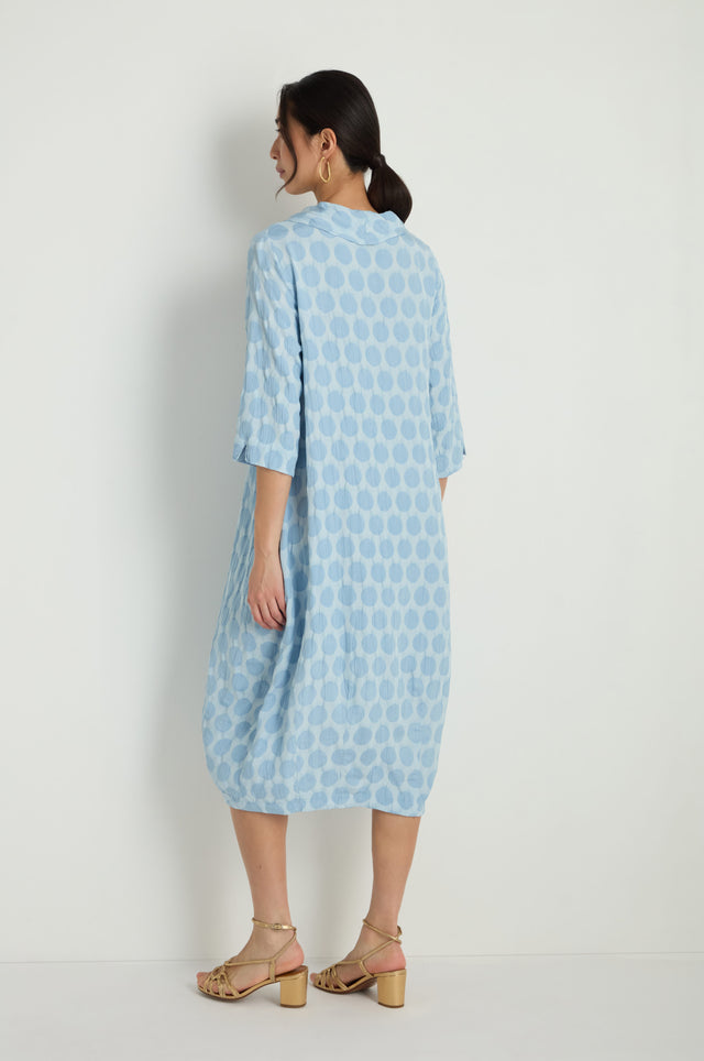A woman smiles and holds the sides of her Spot Textured Jacquard Bubble Dress, a light blue, midi-length garment with a polka dot pattern, while standing against a plain white background.