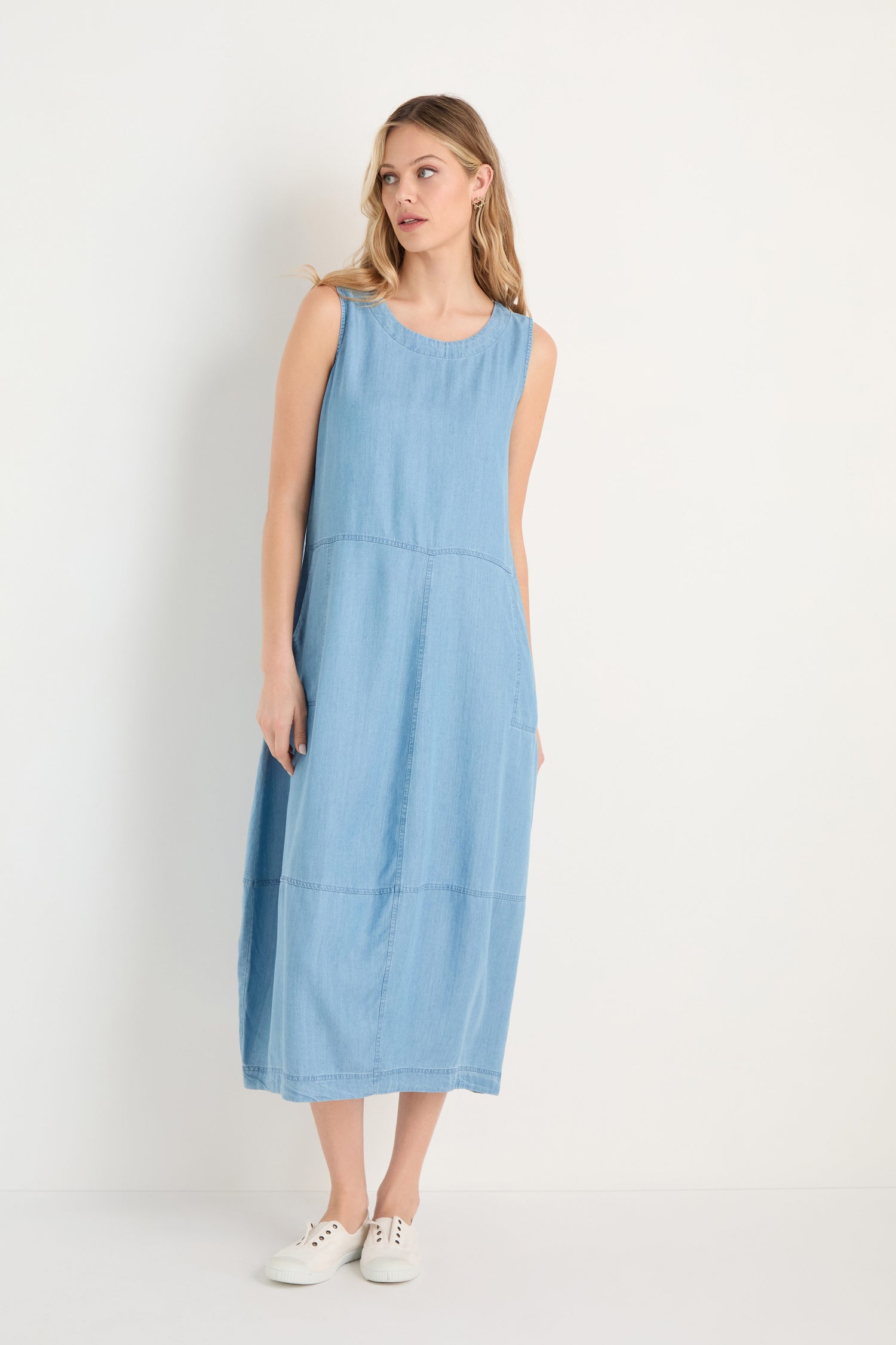 A woman models the Fluid Denim Bubble Dress—a sleeveless, light blue midi dress with a bubble silhouette—paired with white sneakers, set against a plain white background.