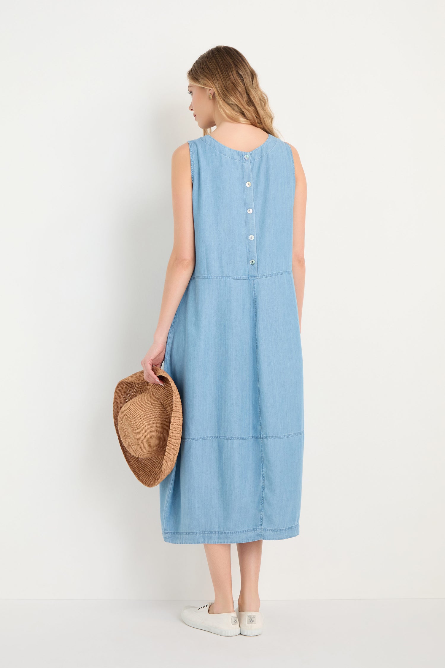 A woman faces away, wearing the Fluid Denim Bubble Dress—a sleeveless, light blue midi with back buttons—holding a straw hat in her right hand and wearing white sneakers.