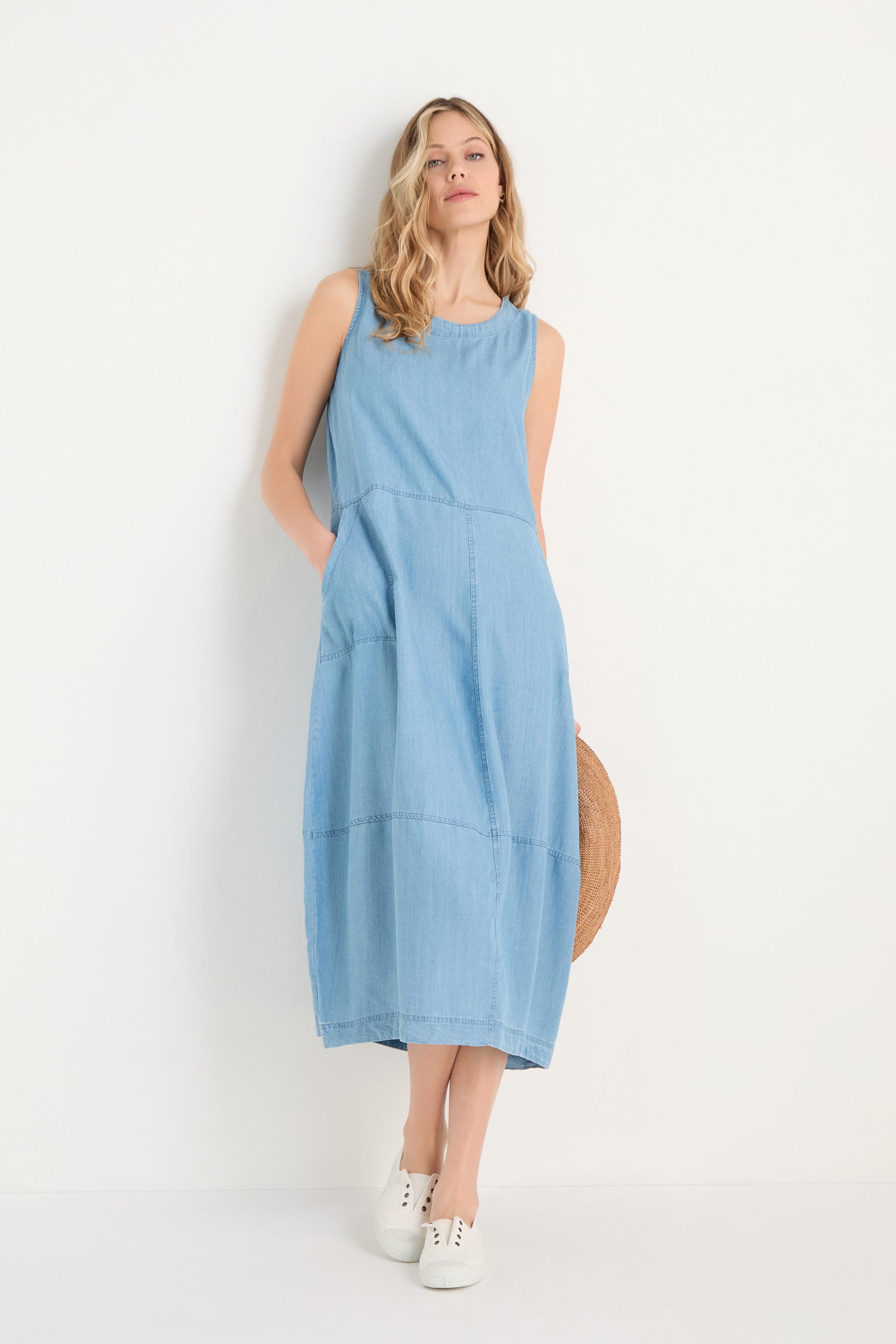 A woman in the Fluid Denim Bubble Dress, a sleeveless midi denim style with a subtle bubble silhouette, pairs it with white sneakers and holds a round straw bag while standing against a plain white background.