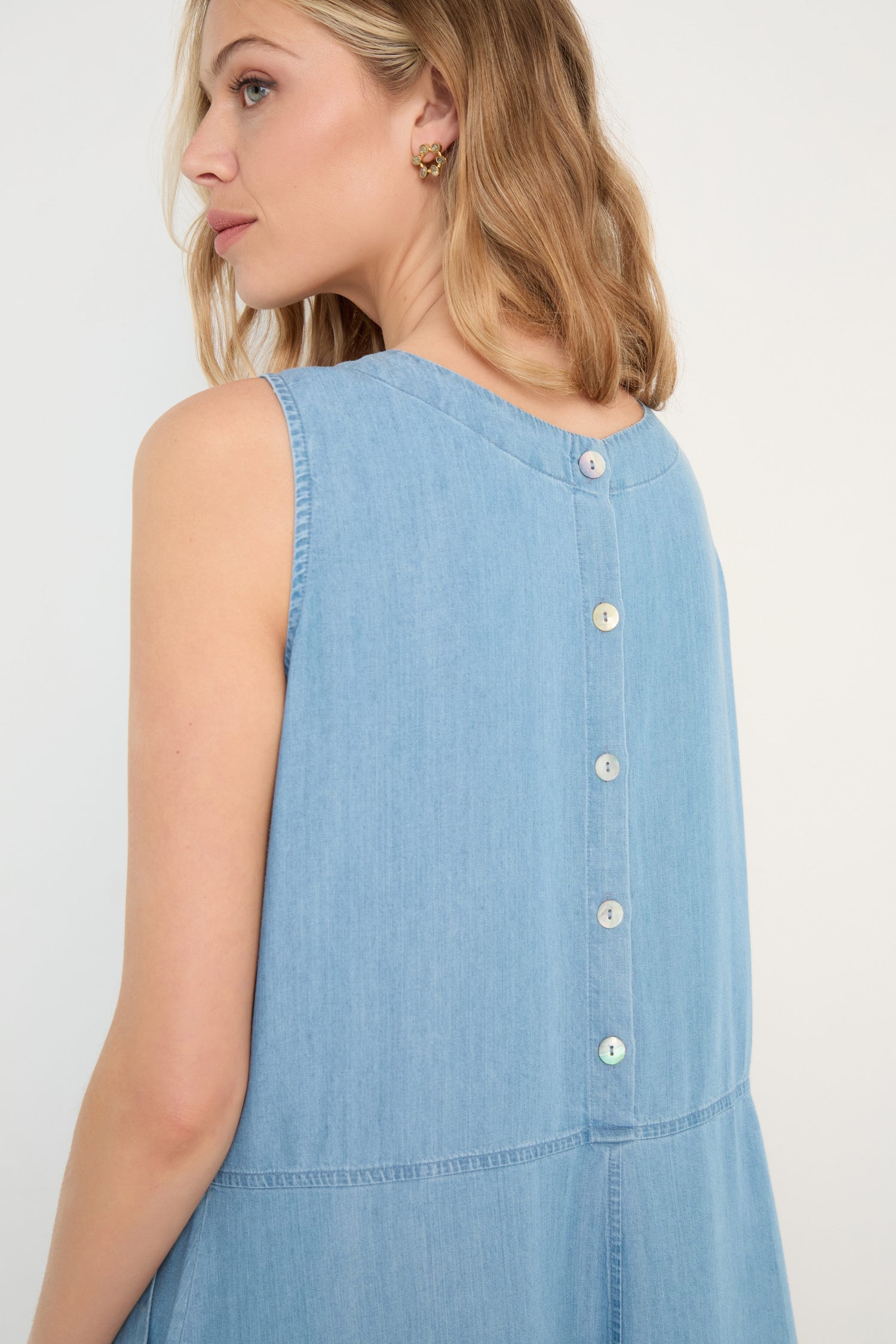 A woman shown from the side and back wears the Fluid Denim Bubble Dress, a sleeveless light blue denim midi dress featuring a bubble silhouette and button-down back.
