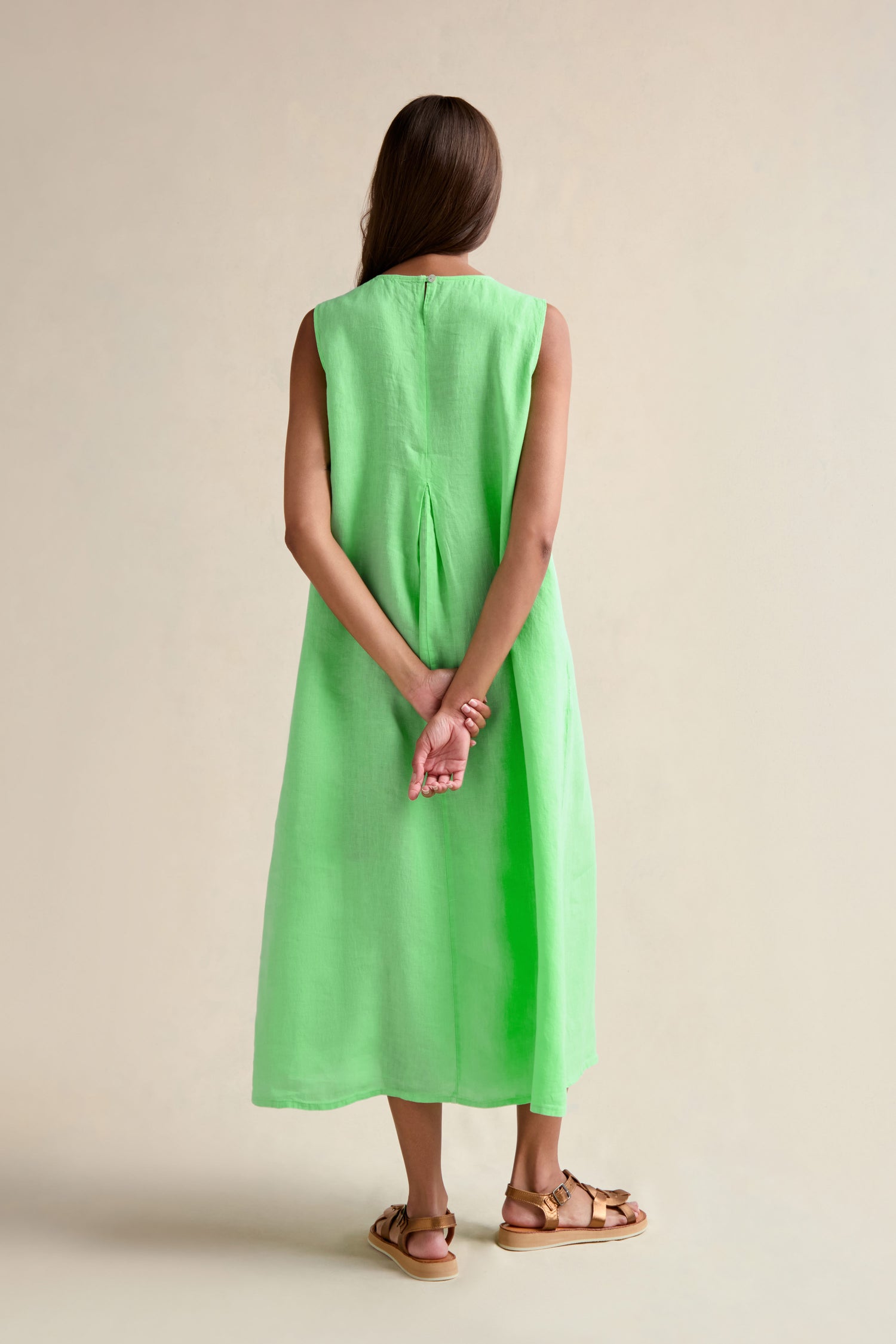 A woman, with her back to the camera, wears the Linen Pleat Front Sleeveless Dress in mint green paired with brown sandals—her hands clasped behind her, showcasing this perfect summer staple.