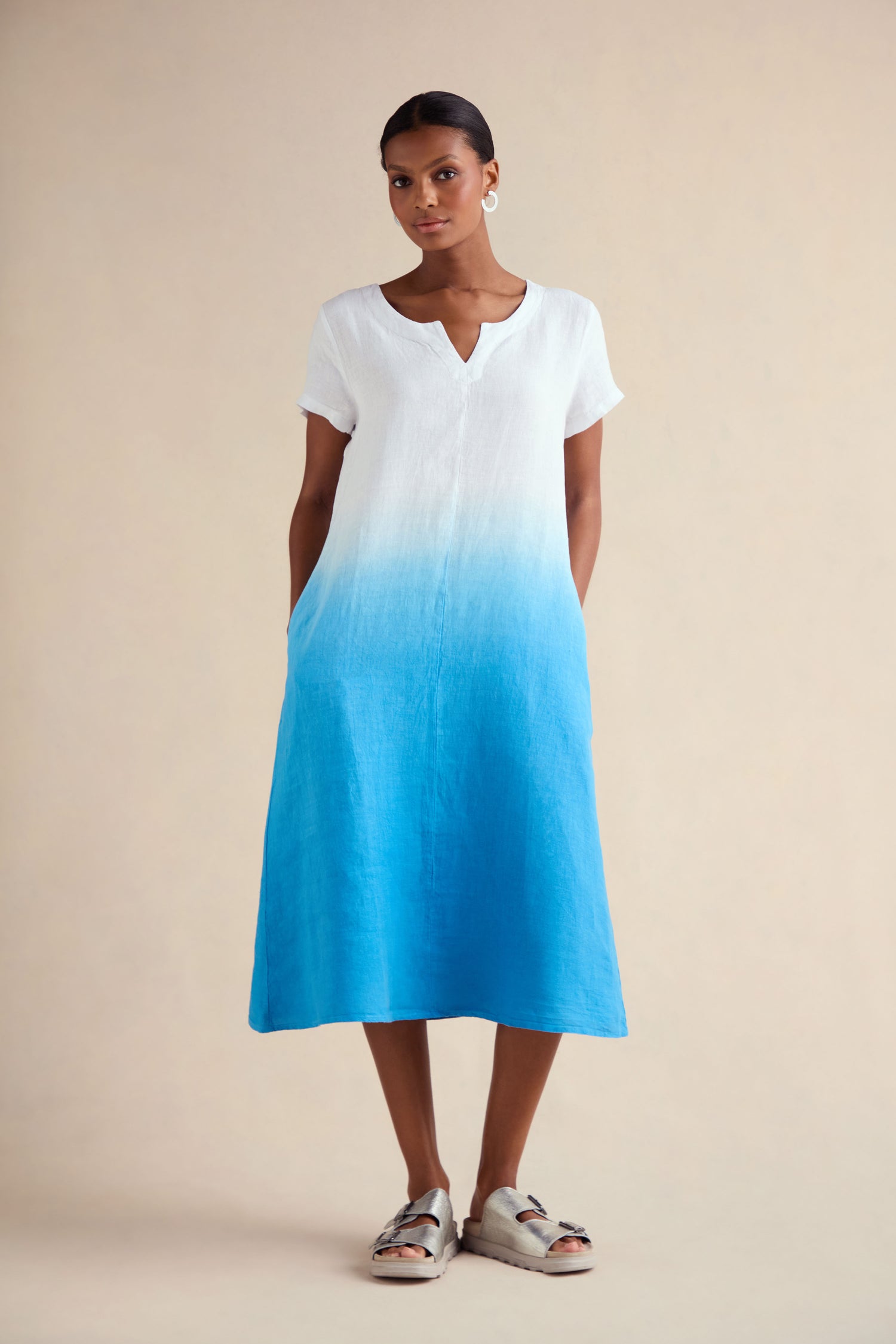 A woman poses against a plain background wearing the Dip Dye Linen Dress—a short-sleeve, knee-length A-line linen dress featuring a white top that fades into blue. She completes the look with sandals.