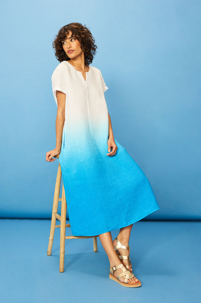 A person with curly hair wears the Dip Dye Linen Dress and gold sandals, sitting on a wooden stool against a blue background.