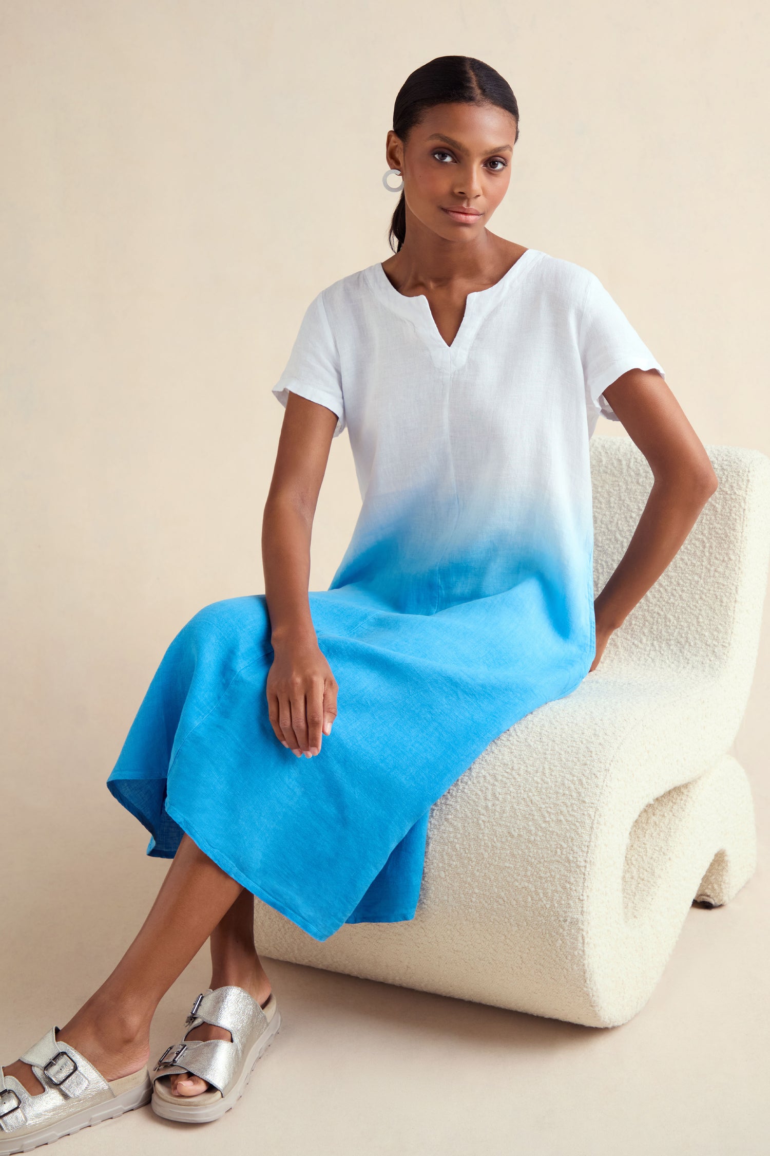 A woman sits on a textured white chair wearing the Dip Dye Linen Dress with silver sandals against a neutral background.