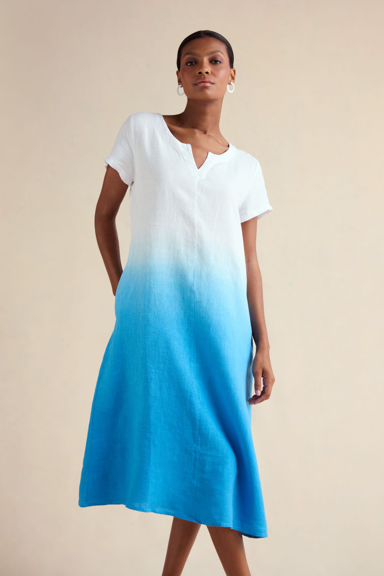 A woman wears the Dip Dye Linen Dress, featuring a short-sleeve V-neck design with a white top blending into a bright blue ombre A-line skirt, standing against a plain light background.