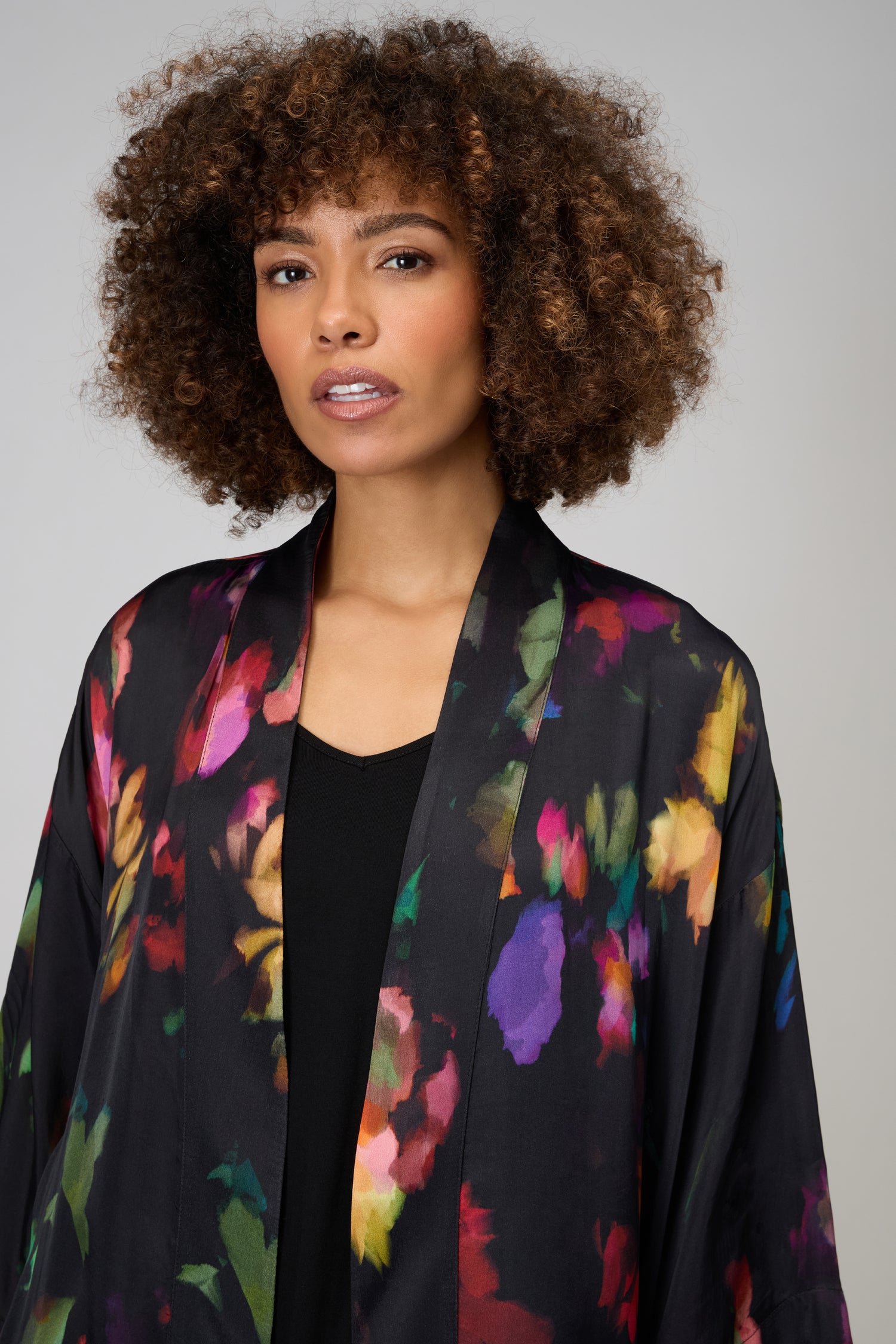 A person with curly hair wears a black top layered with the Winter Floral Kimono, featuring dark painterly florals, standing against a plain light background.
